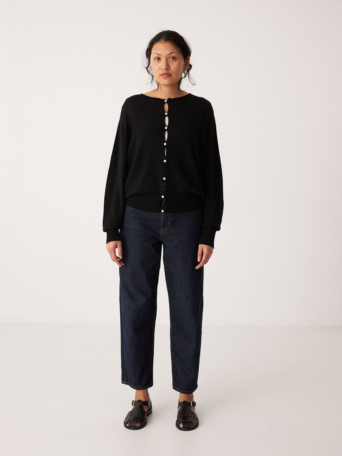 A woman stands against a plain background wearing the Francie Anni Cardigan in black, paired with dark blue jeans and black sandals.