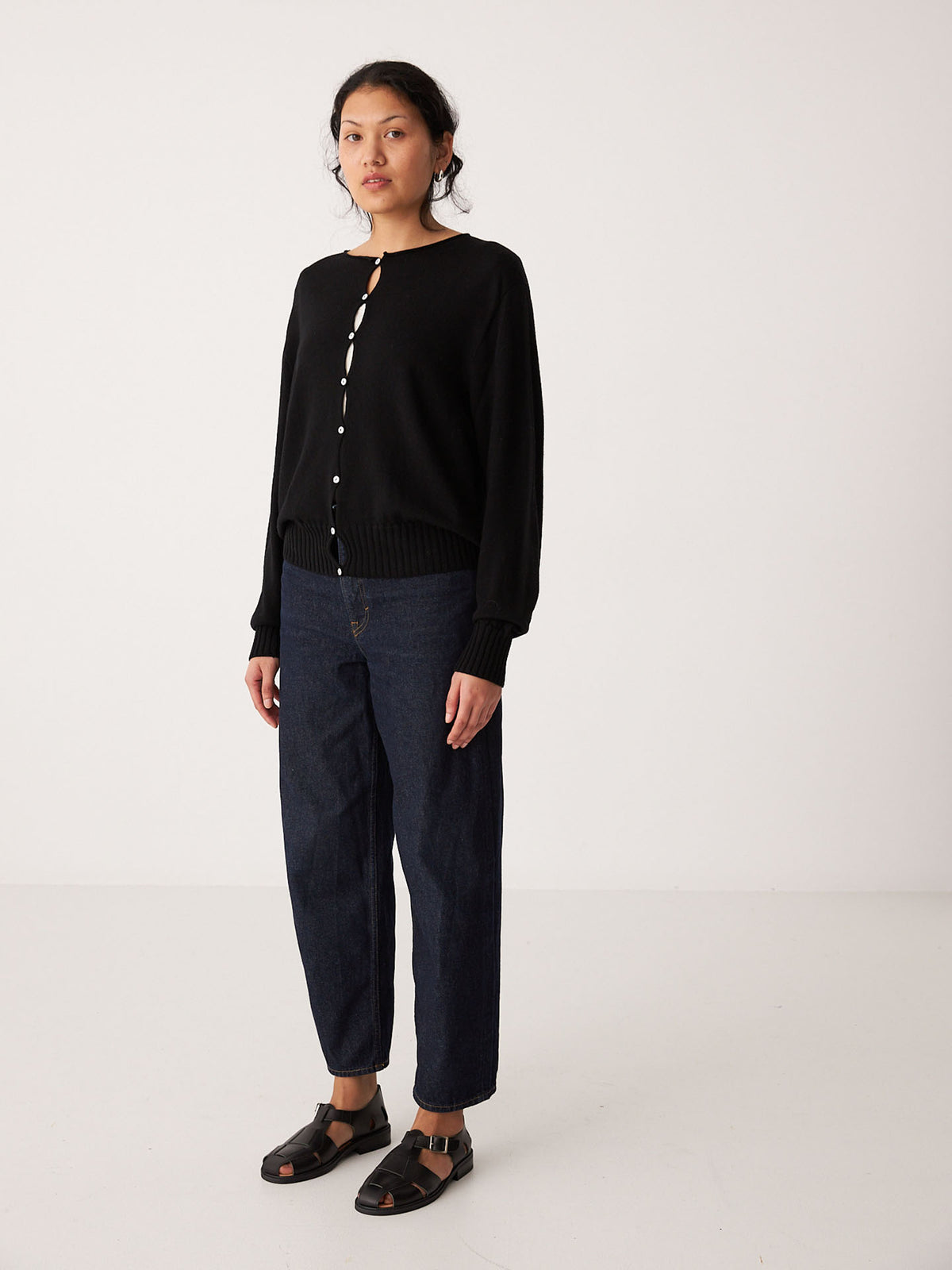 A petite person stands in a plain room wearing Francie’s Anni Cardigan in black, styled with dark blue jeans and black sandals.