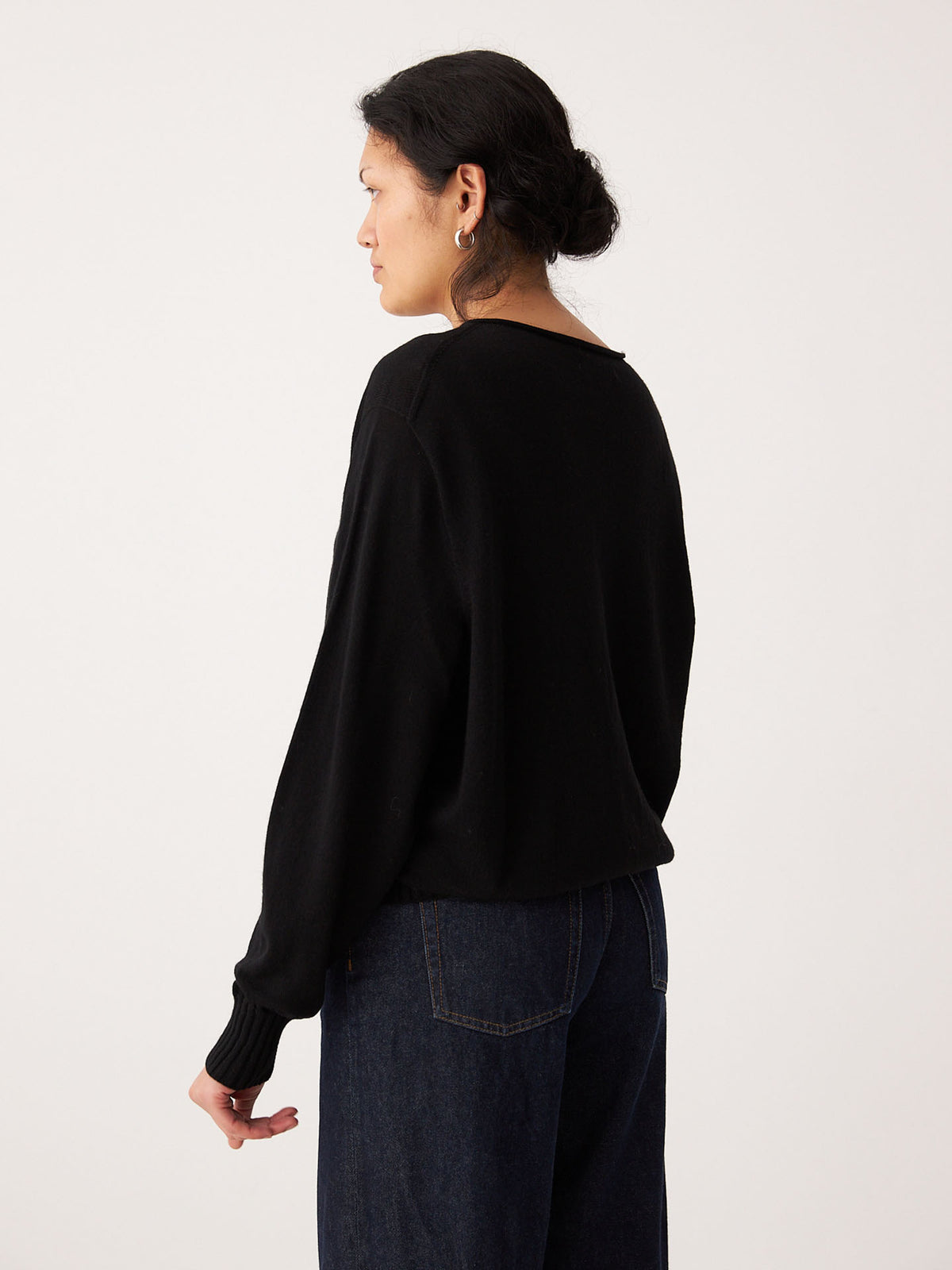 A petite person with dark hair in a low bun wears the Francie Anni Cardigan in Black and relaxed fit dark blue jeans, standing facing away against a plain light background.