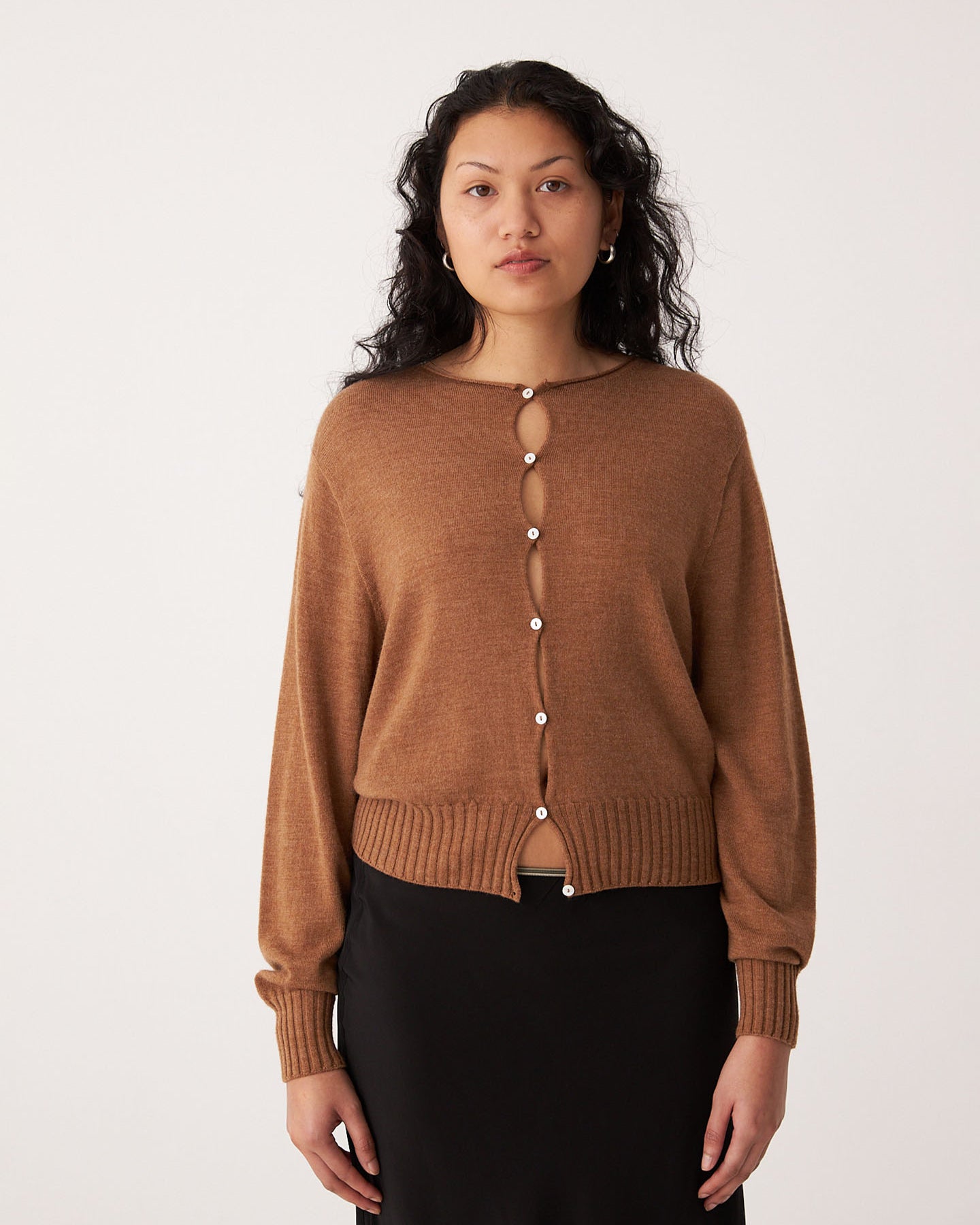 A person with long, wavy hair wears Francie's Anni Cardigan in Brown Sugar and a black skirt, standing against a plain white background.