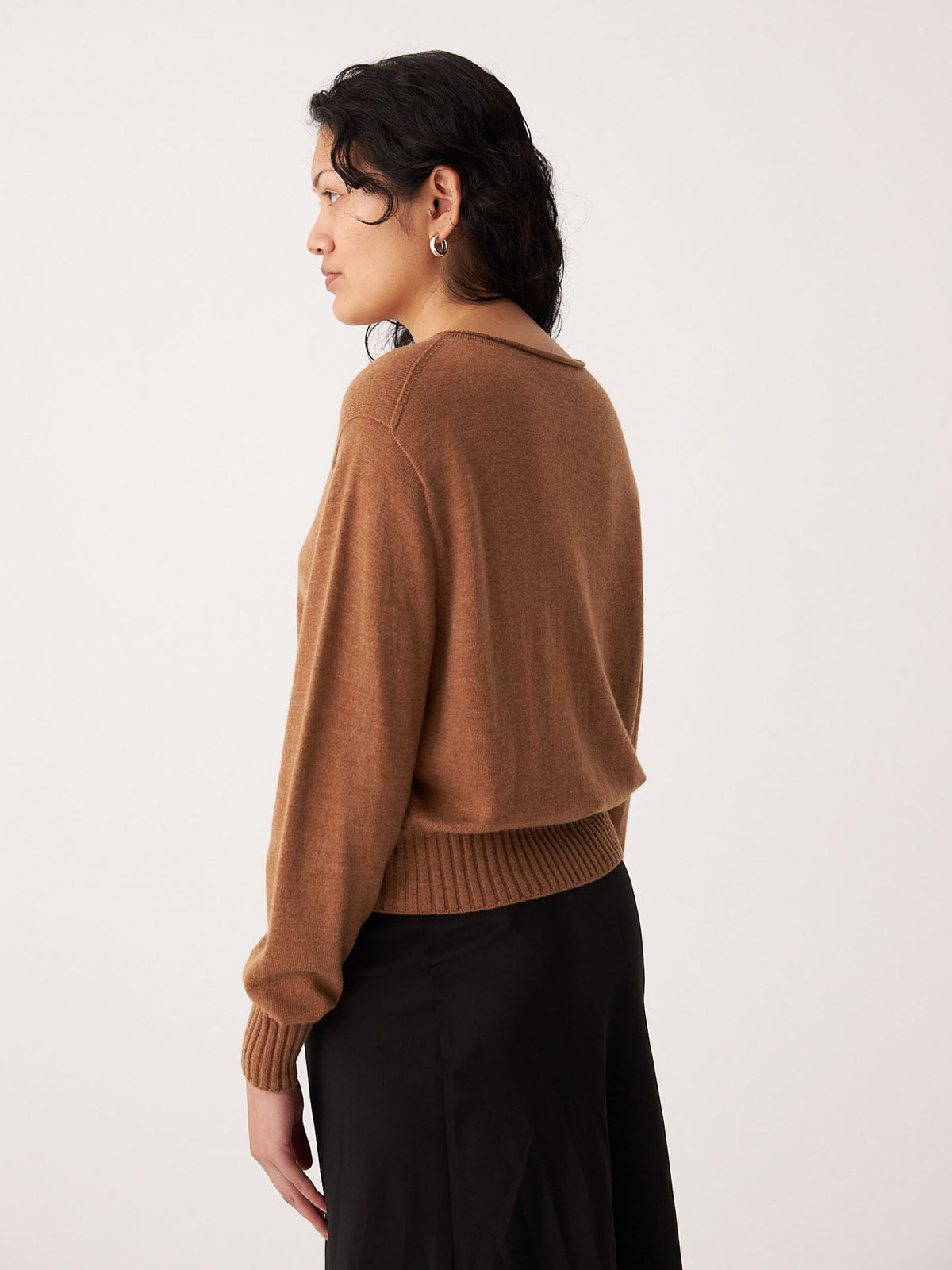 A person with dark hair, wearing the Francie Anni Cardigan in Brown Sugar and a black skirt, stands sideways against a plain background.