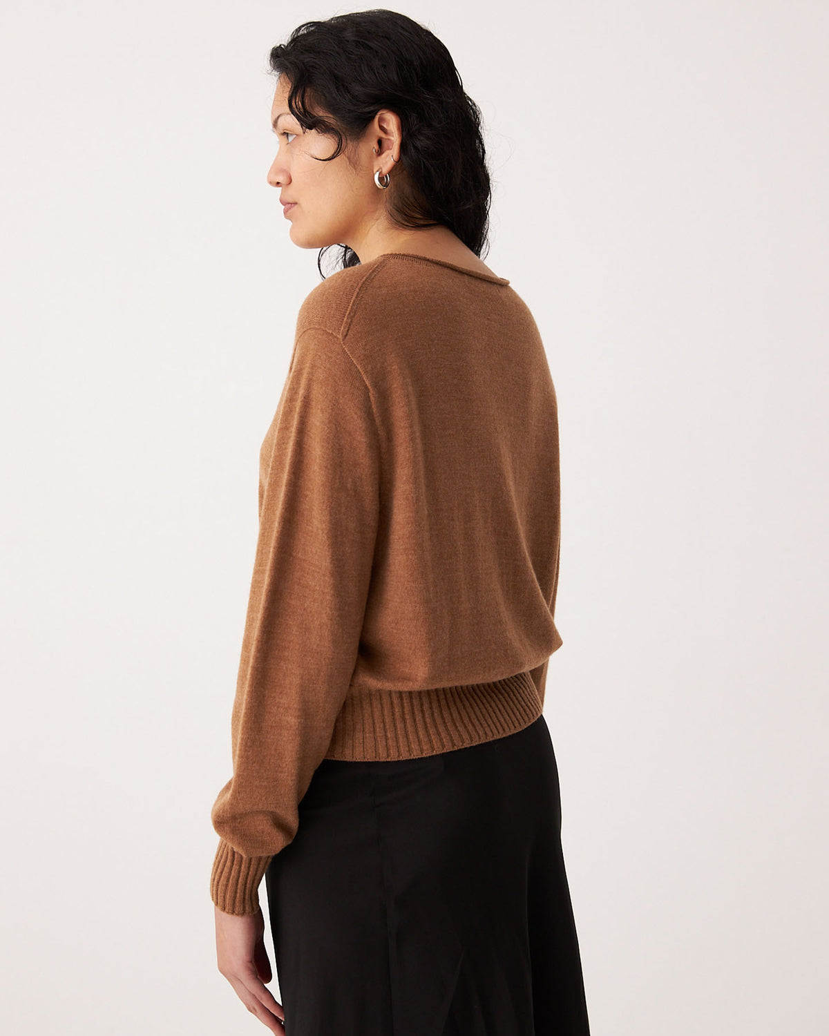 A person with dark hair, wearing the Francie Anni Cardigan in Brown Sugar and a black skirt, stands sideways against a plain background.