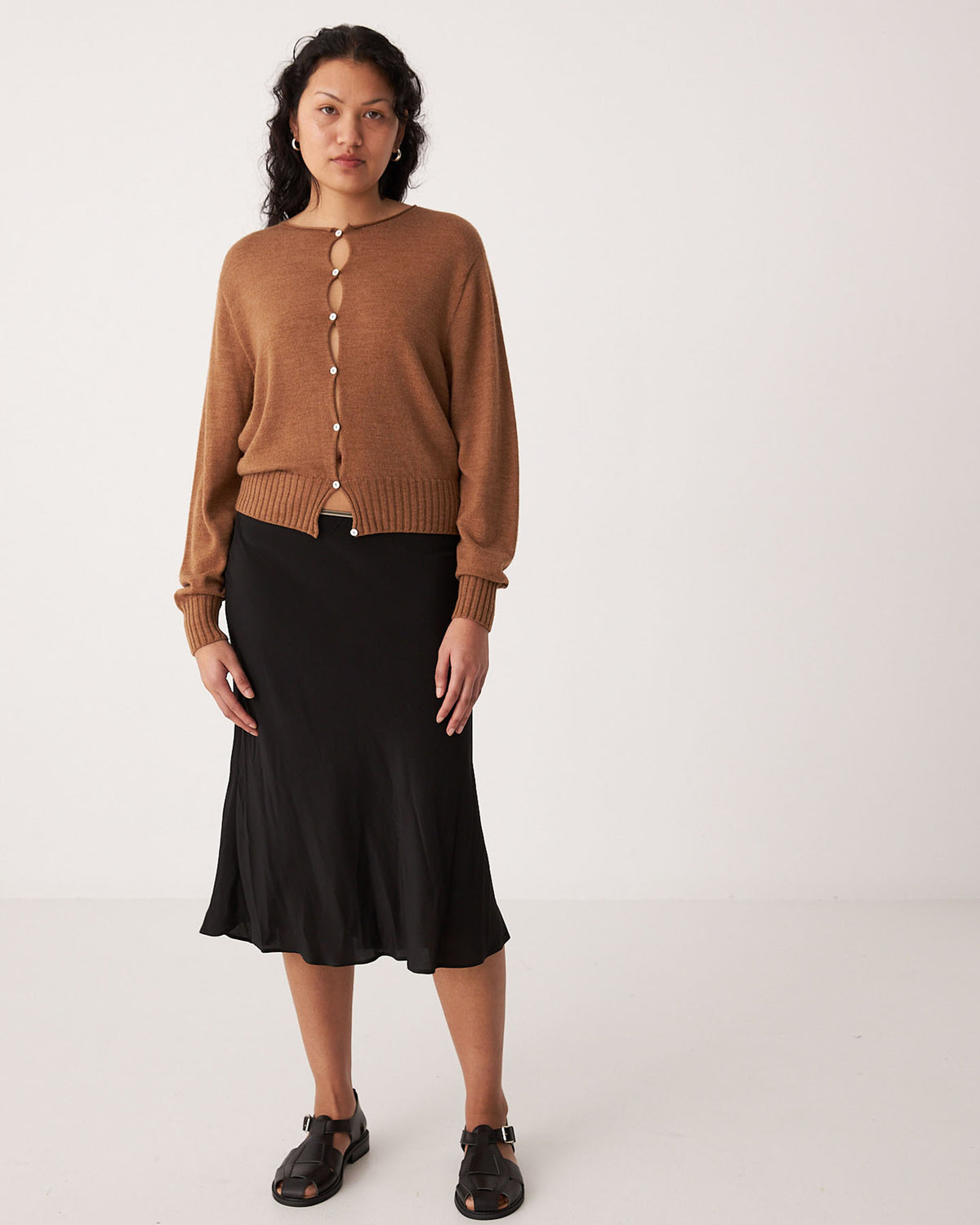 A petite woman stands in a plain room wearing the Francie Anni Cardigan in Brown Sugar, paired with a black midi skirt and black sandals.