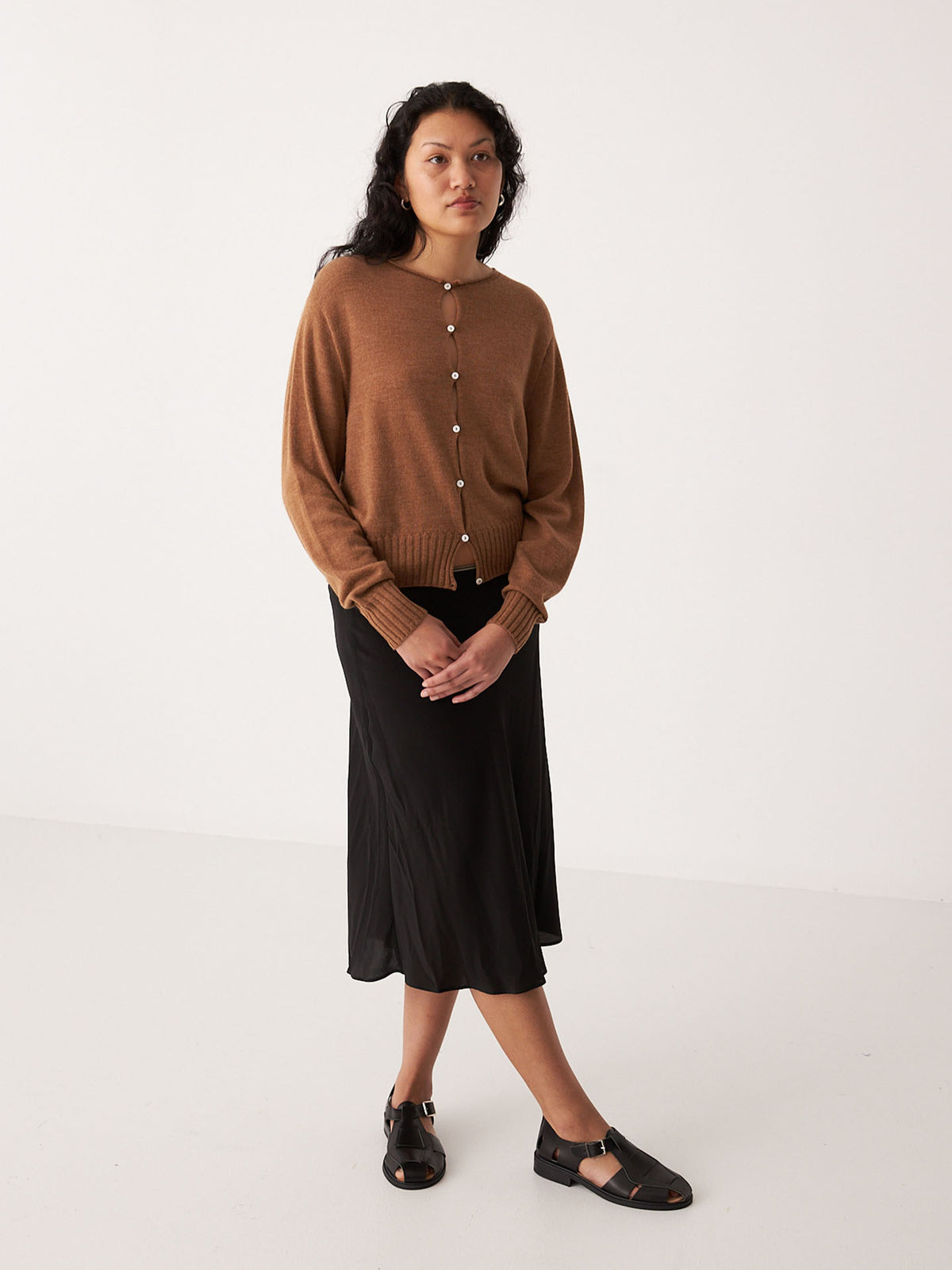 A petite woman stands against a plain background wearing the Anni Cardigan in Brown Sugar by Francie, paired with a black midi skirt and black sandals.