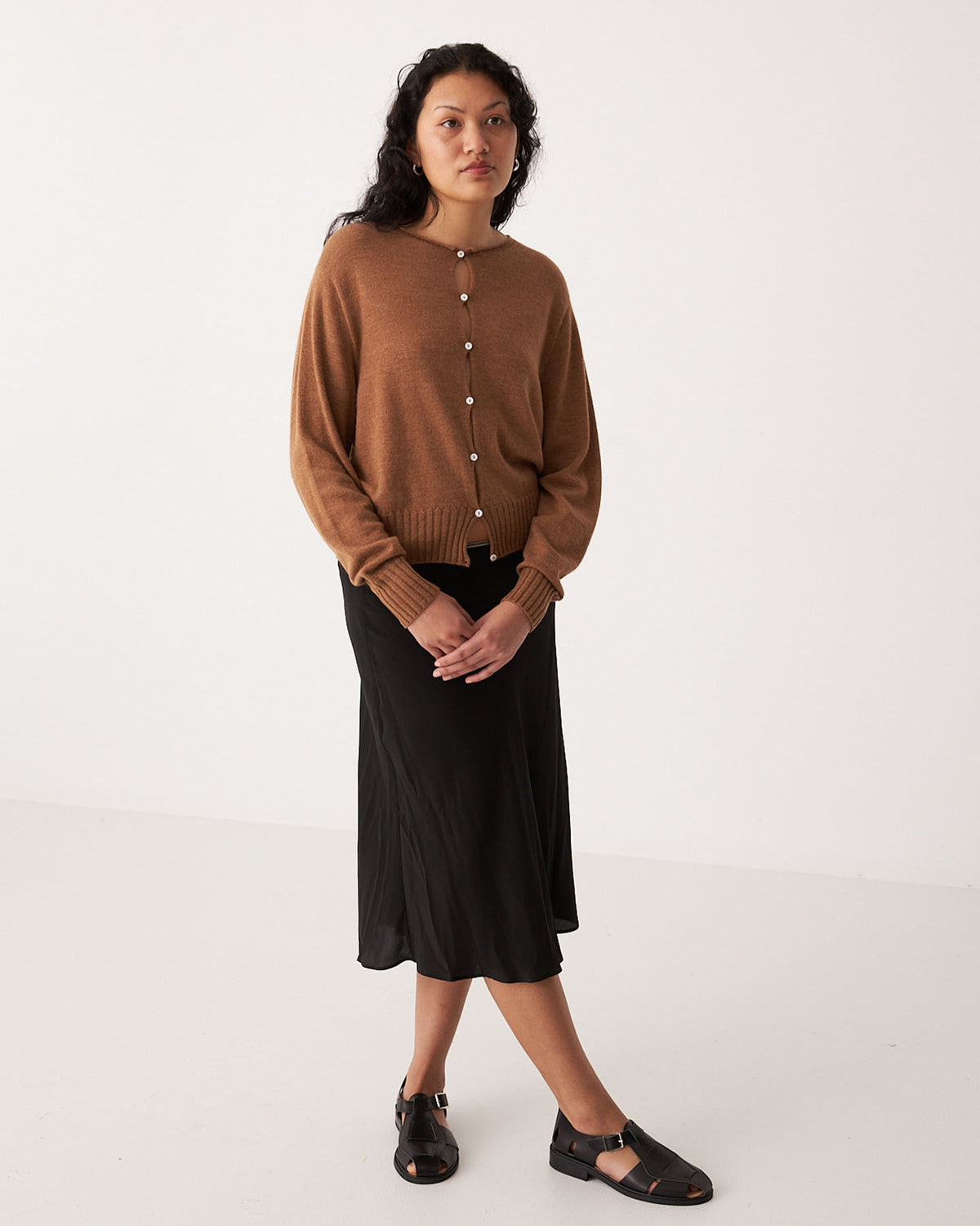 A petite woman stands against a plain background wearing the Anni Cardigan in Brown Sugar by Francie, paired with a black midi skirt and black sandals.