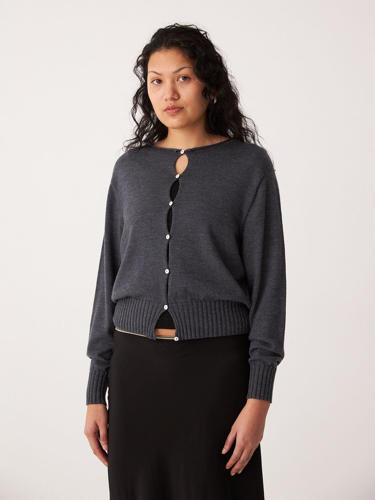 A woman wears the Francie Anni Cardigan in Shadow Grey, a relaxed fit with a keyhole opening, paired with a black skirt against a plain light background.