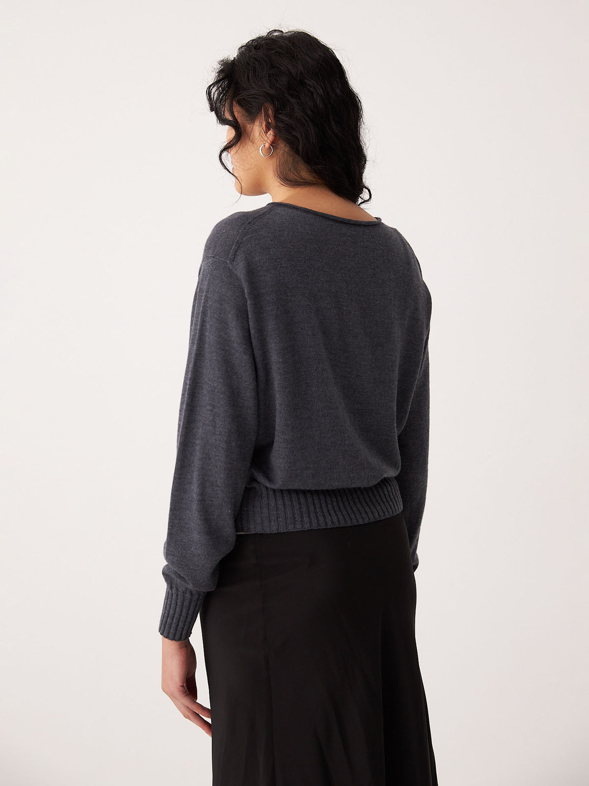 A woman with dark hair stands facing away, wearing the Francie Anni Cardigan in Shadow Grey and a black skirt against a plain light background.