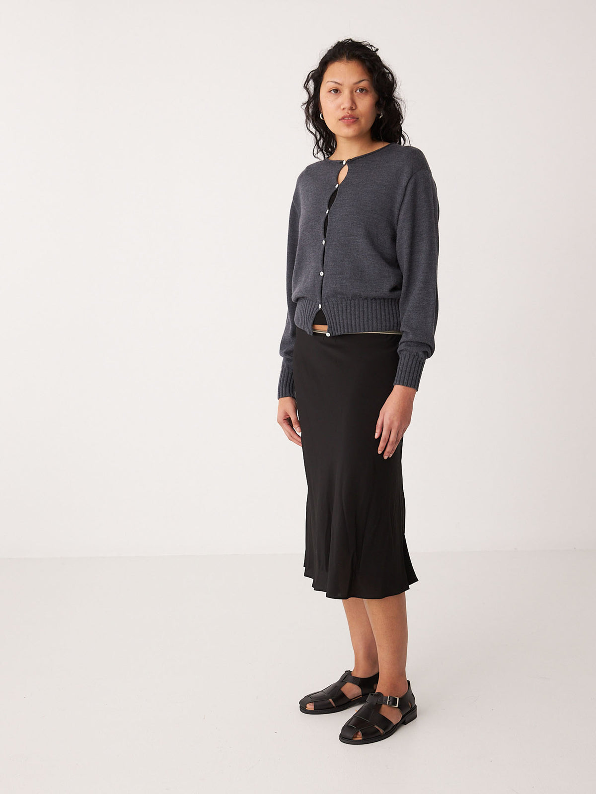 A woman stands in a minimalist setting wearing Francie’s Anni Cardigan in Shadow Grey, paired with a black midi skirt and black sandals.