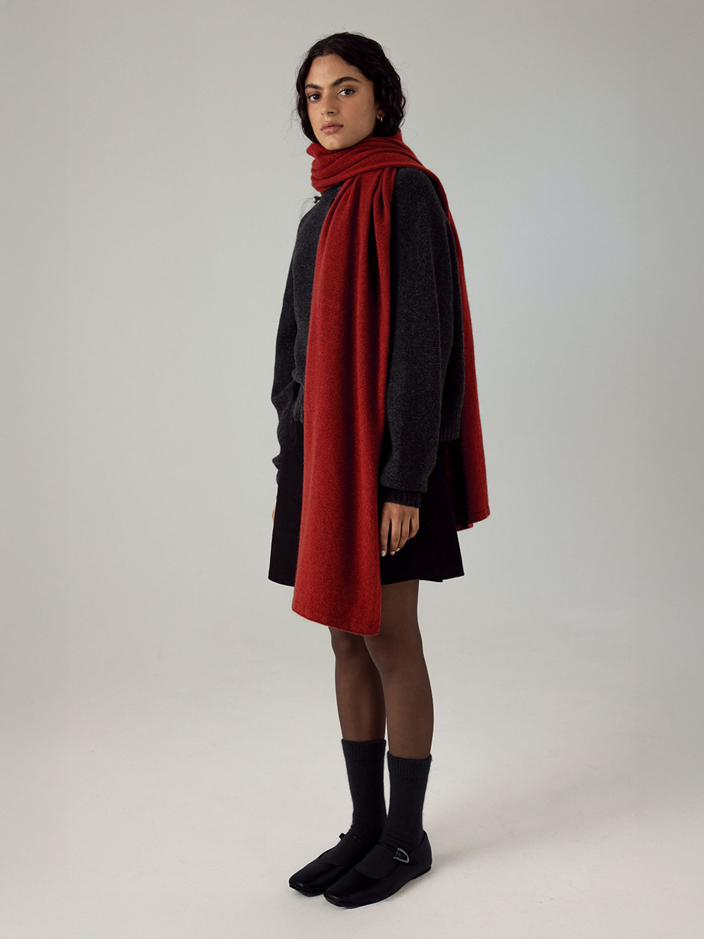A person stands against a plain light background beside an extra wide yoga mat, wearing a dark sweater, black skirt, black shoes with socks, and accessorized with the Francie Cloud Scarf – Poppy.