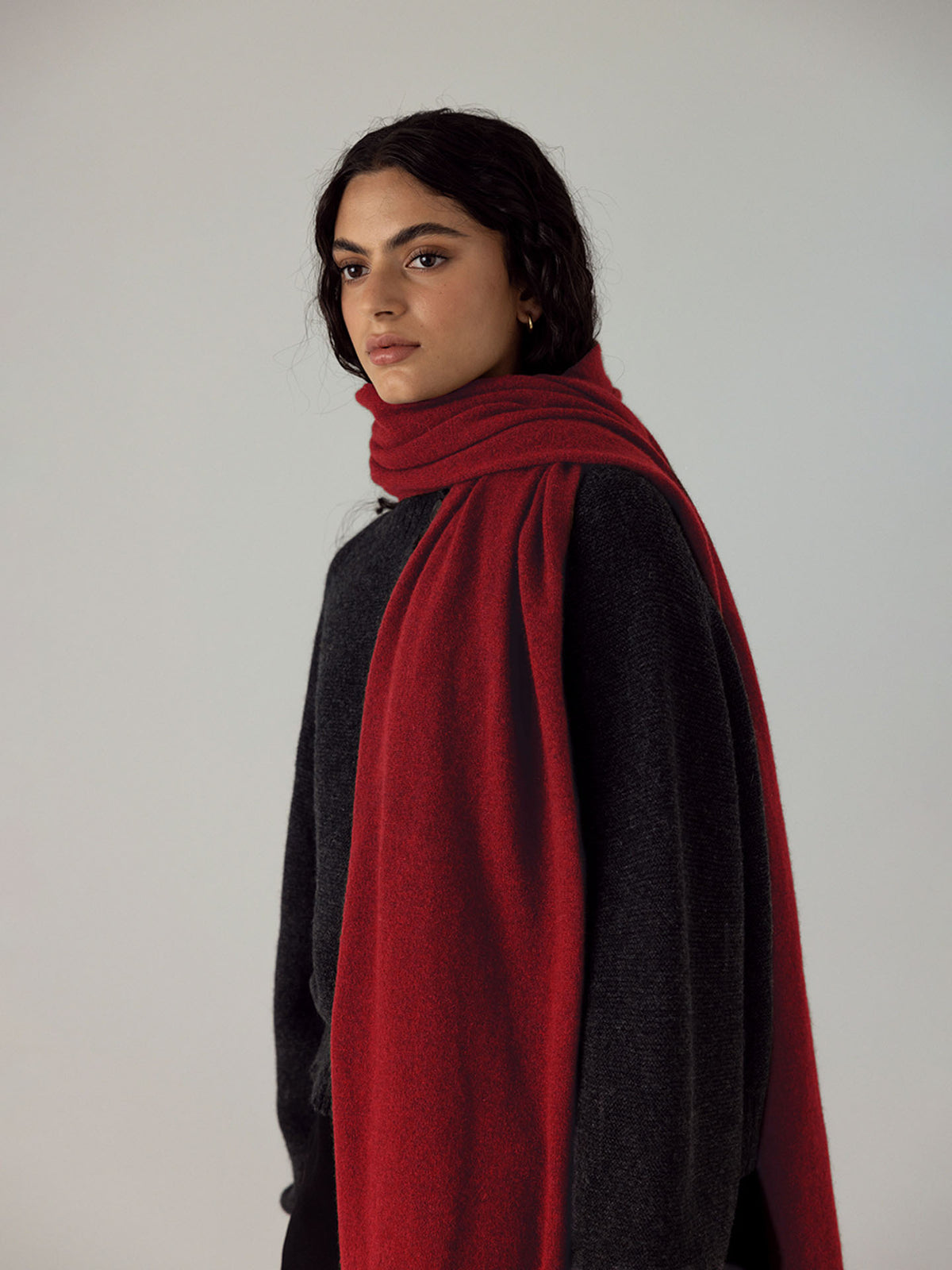 A person with long dark hair wears a dark sweater and the Francie Cloud Scarf in Poppy, draped over their shoulders, standing by an extra wide yoga mat against a plain light background.