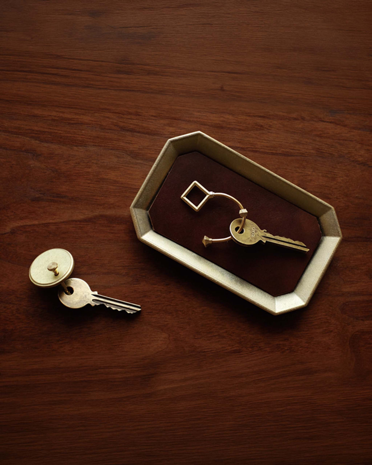 Two gold keys, one on a key ring in a tray and one attached to a round key tag, placed on a wooden surface.