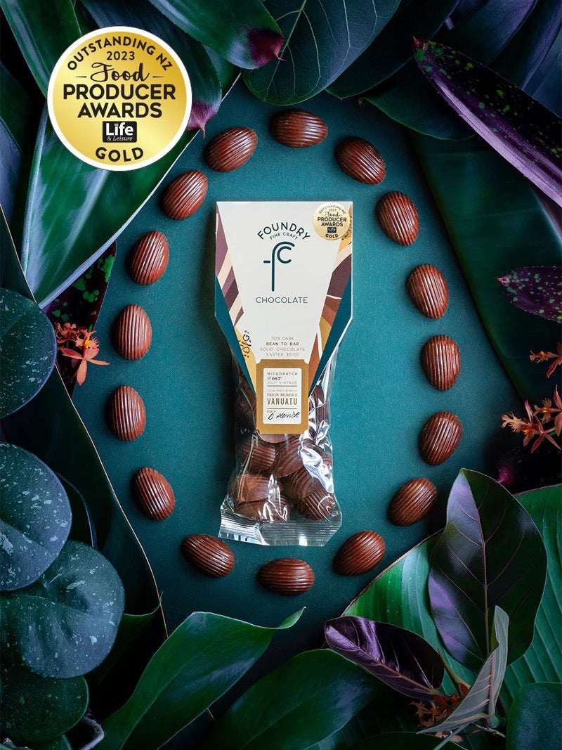 Package of Mini Chocolate Eggs - Vanuatu 70% Foundry Chocolate amid scattered hazelnuts and green foliage, with an &quot;outstanding nz food producer awards 2023 gold&quot; badge.