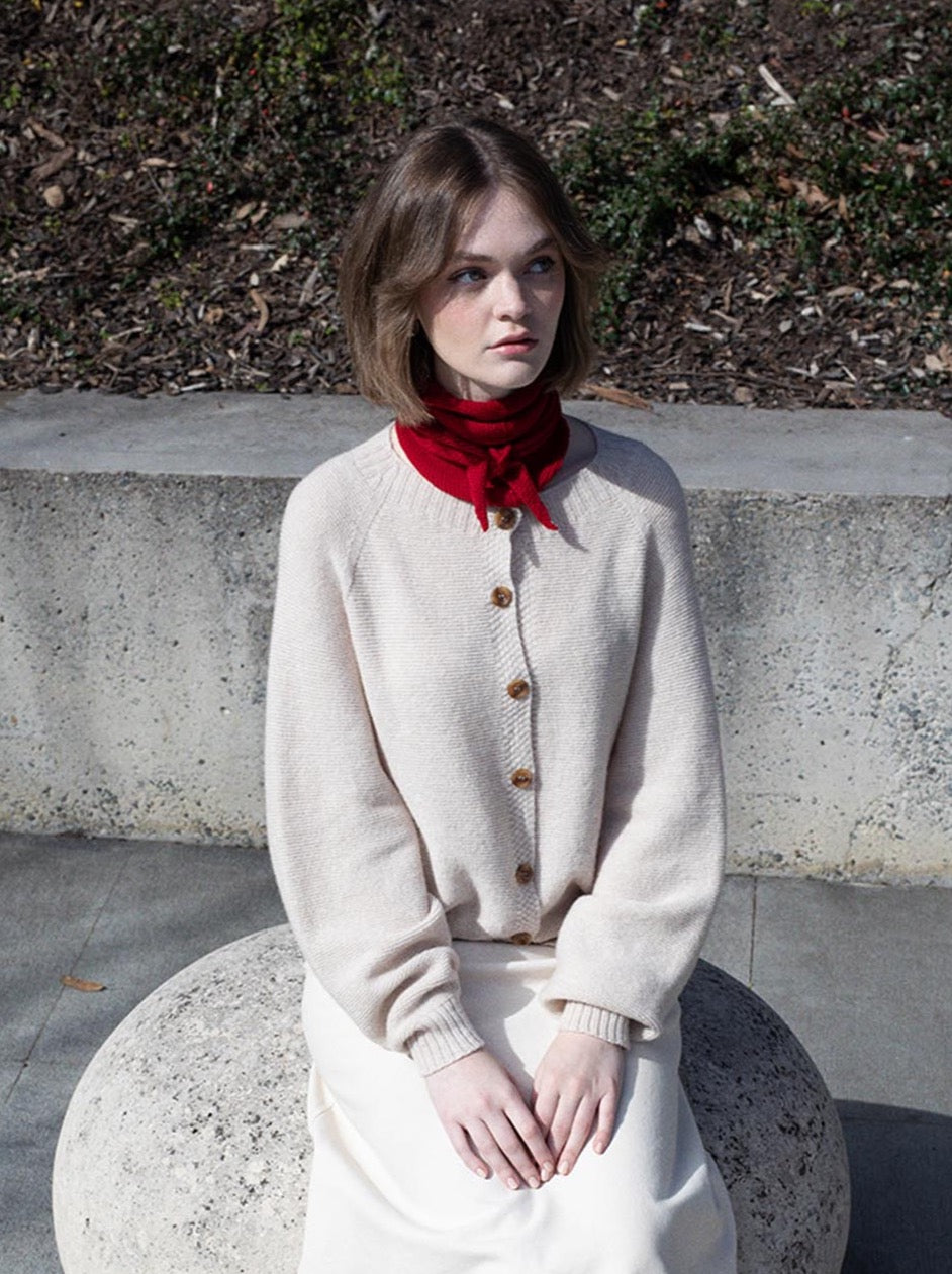 A woman sitting on a rock, wrapped in an ethically made Francie Merino wool Daisy Scarf – Crimson.