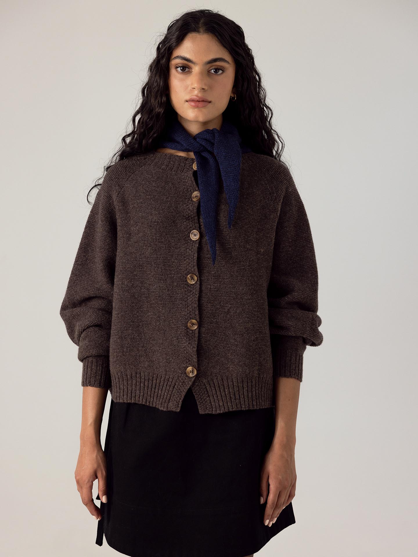 The model is wearing a brown cardigan, made from 100% Extra-fine Merino Wool, and a black skirt. She is also wearing the Daisy Scarf in Midnight from Francie.