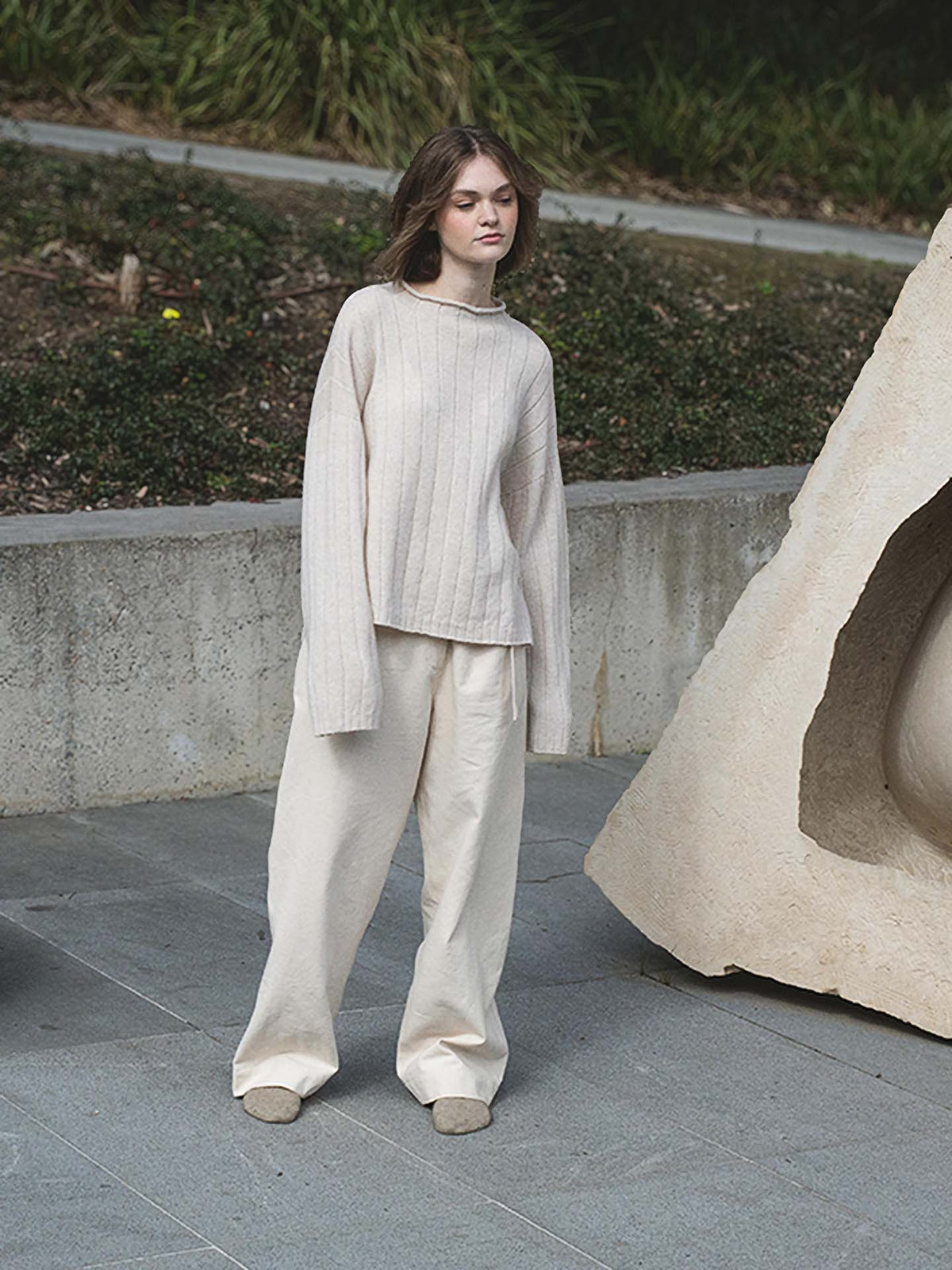 A woman standing in front of a sculpture wearing an oversized Francie Echo Knit – Creme sweater made from 100% Merino Wool and wide leg pants.