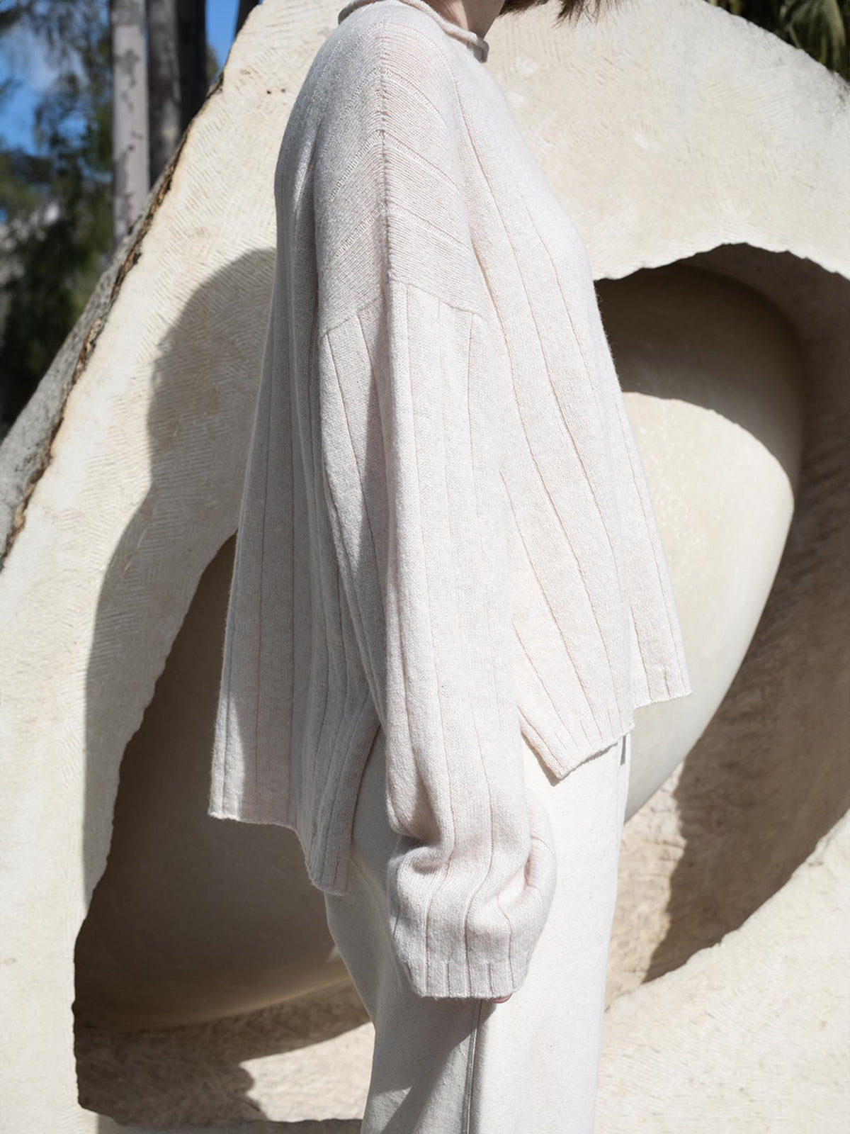 A woman wearing an oversized white sweater and pants, ethically knitted from Italian air-spun merino, in front of a sculpture.