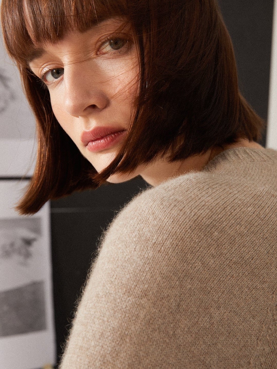 Young woman with bob haircut, wearing a fitted beige sweater, looking over her shoulder in a room with black and white photos on the wall wearing the Francie Nimbus Raglan Knit – Natural.