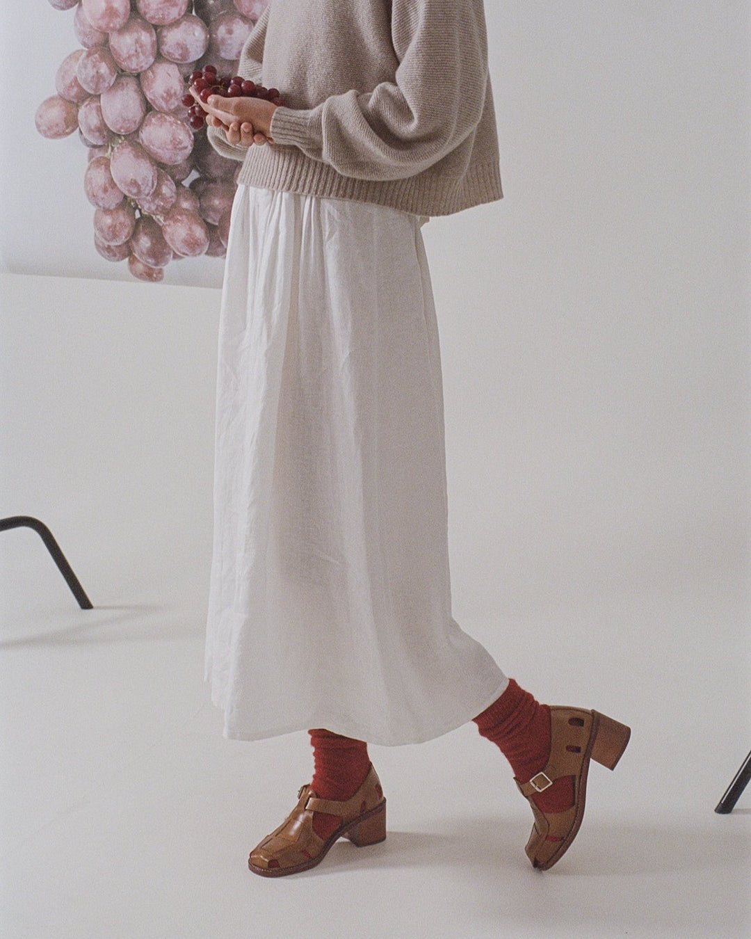 Person in a beige sweater and white skirt holding grapes, with Francie Possum Merino Socks – Crimson and brown shoes (refer to size chart for details), standing in a studio setting.