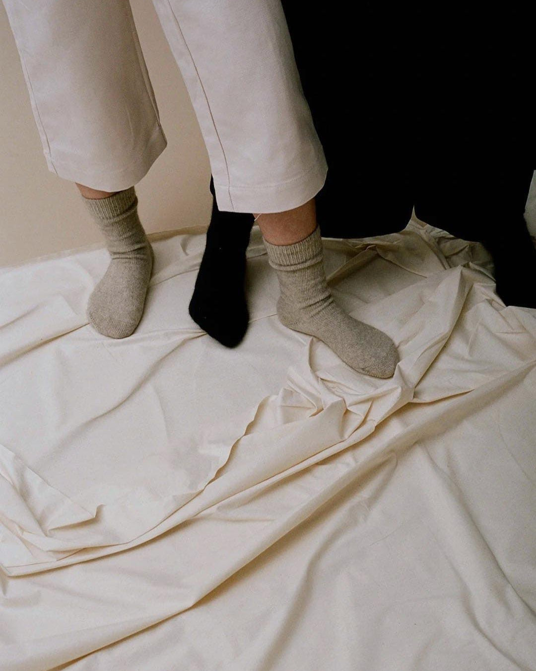 A person standing on a wrinkled beige sheet wearing one black shoe and one Francie Possum Merino Sock – Natural.