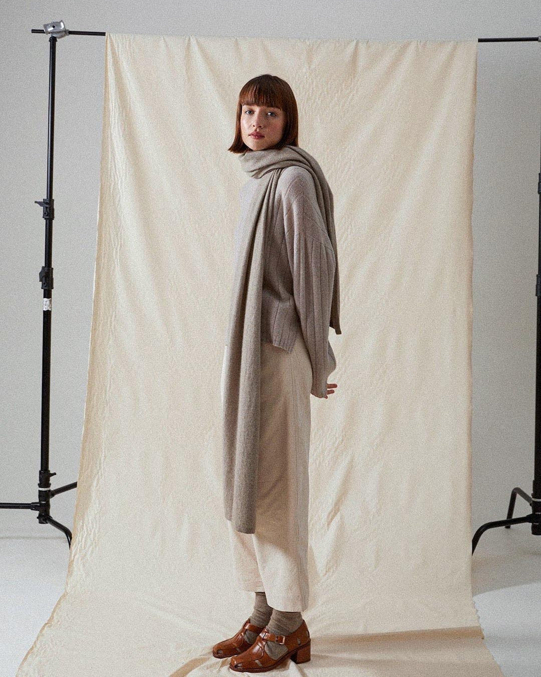 A person stands in front of a neutral-toned backdrop wearing layered, loose-fitting clothes in shades of beige and brown, and brown shoes with New Zealand merino wool socks. The individual is adopting Francie Possum Merino Socks – Natural.