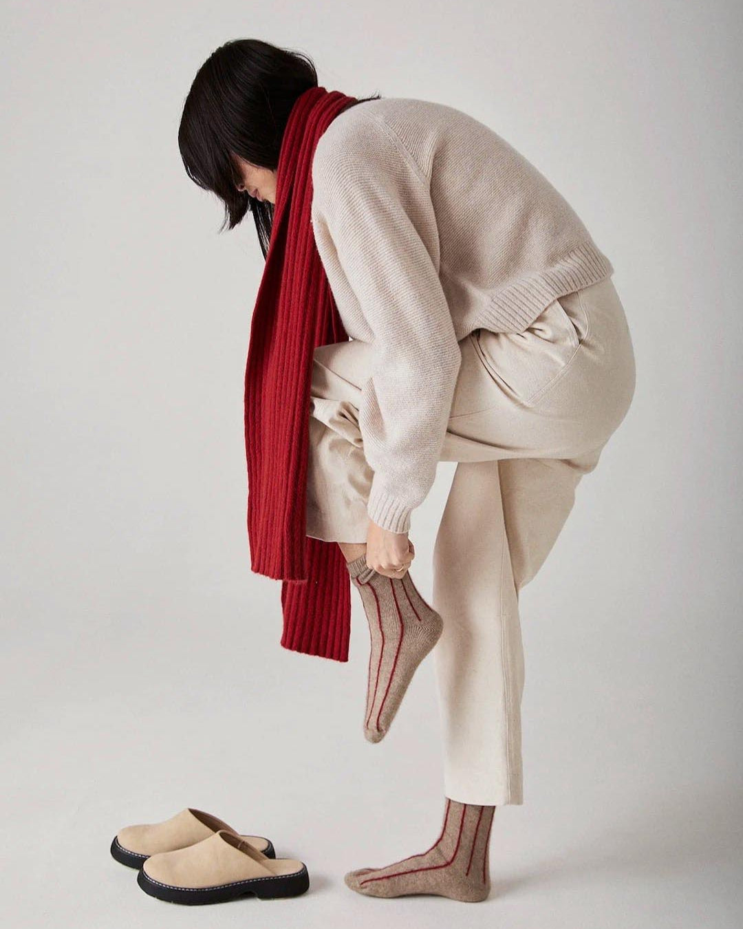 A person in cream-colored pants and a matching sweater is bending over to put on a brown Francie Possum Merino Sock – Natural & Poppy Stripe, with one foot placed on the other knee. They are also wearing a red scarf.