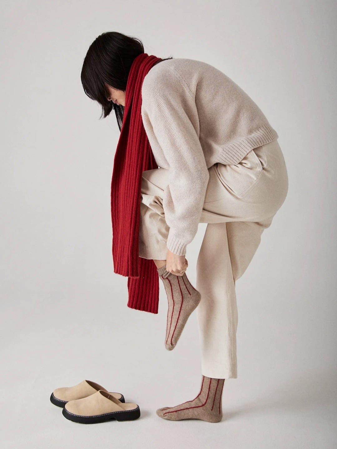 A person in cream-colored pants and a matching sweater is bending over to put on a brown Francie Possum Merino Sock – Natural &amp; Poppy Stripe, with one foot placed on the other knee. They are also wearing a red scarf.
