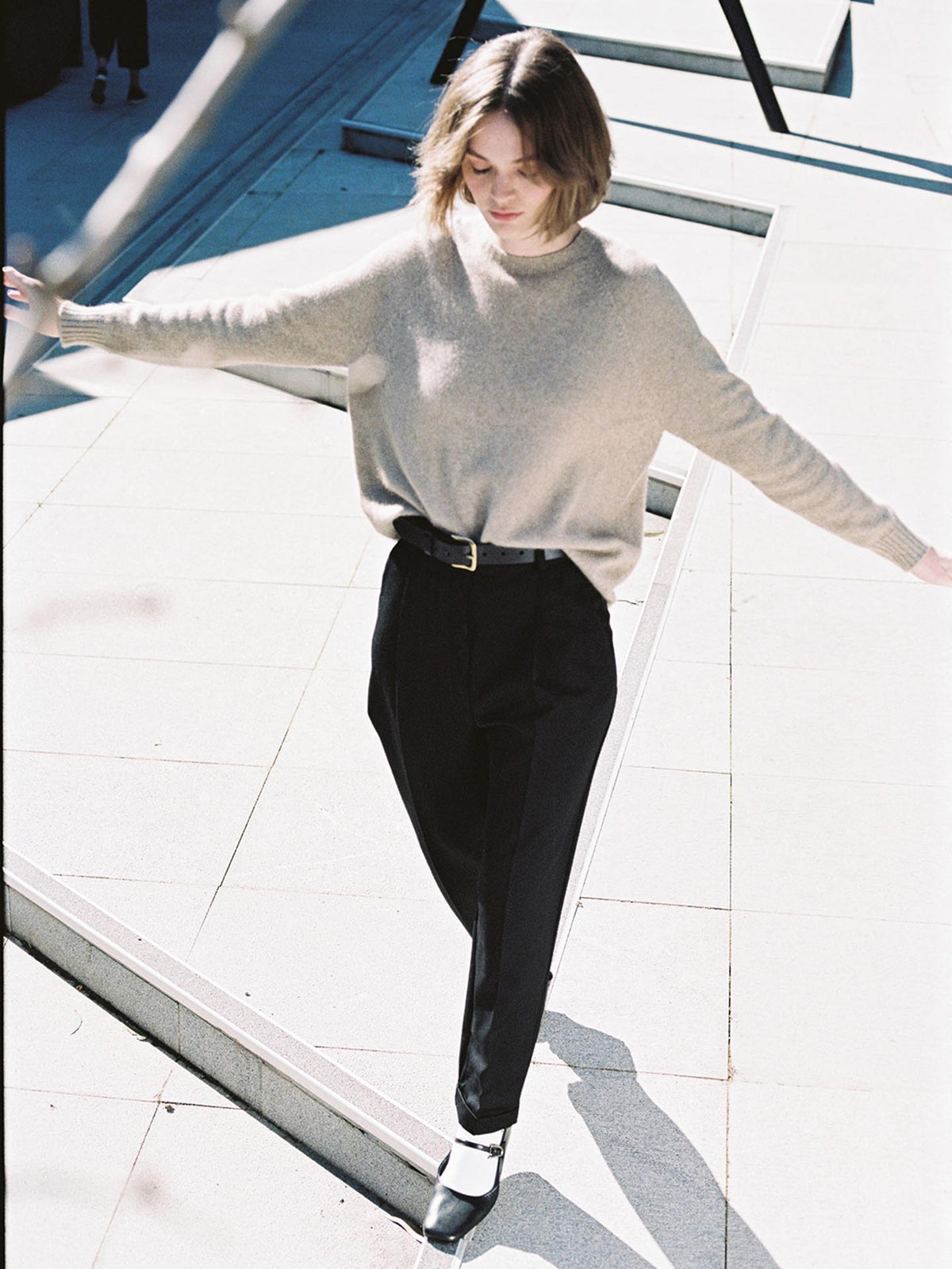 A woman in a Francie Nimbus Raglan Knit – Natural sweater and black trousers walks on a sunny outdoor walkway, arms outstretched for balance.