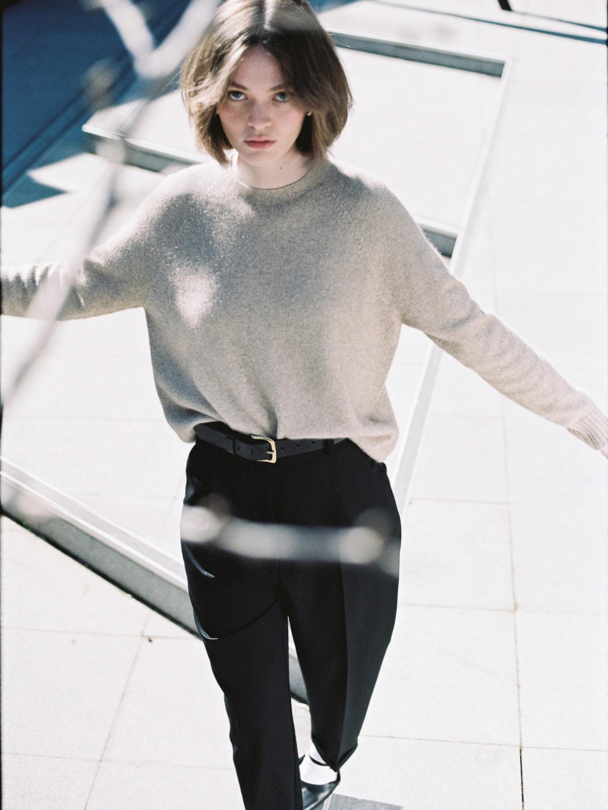 A young woman in a relaxed fit Nimbus Raglan Knit – Natural sweater from Francie and black trousers stands on a sunlit staircase, her hands extended slightly to her sides.