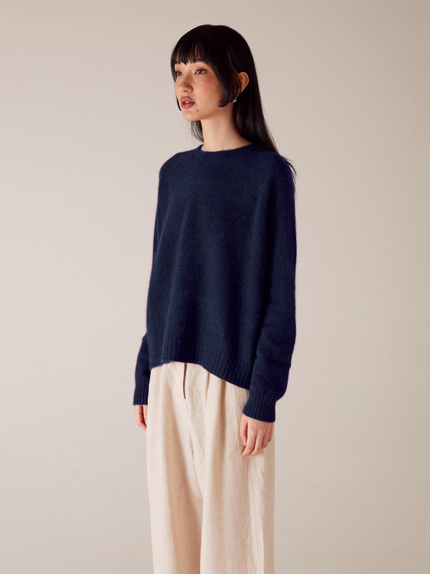 A petite woman with dark hair wearing a Francie Nimbus Raglan Knit – Midnight sweater and beige trousers standing against a light beige background.