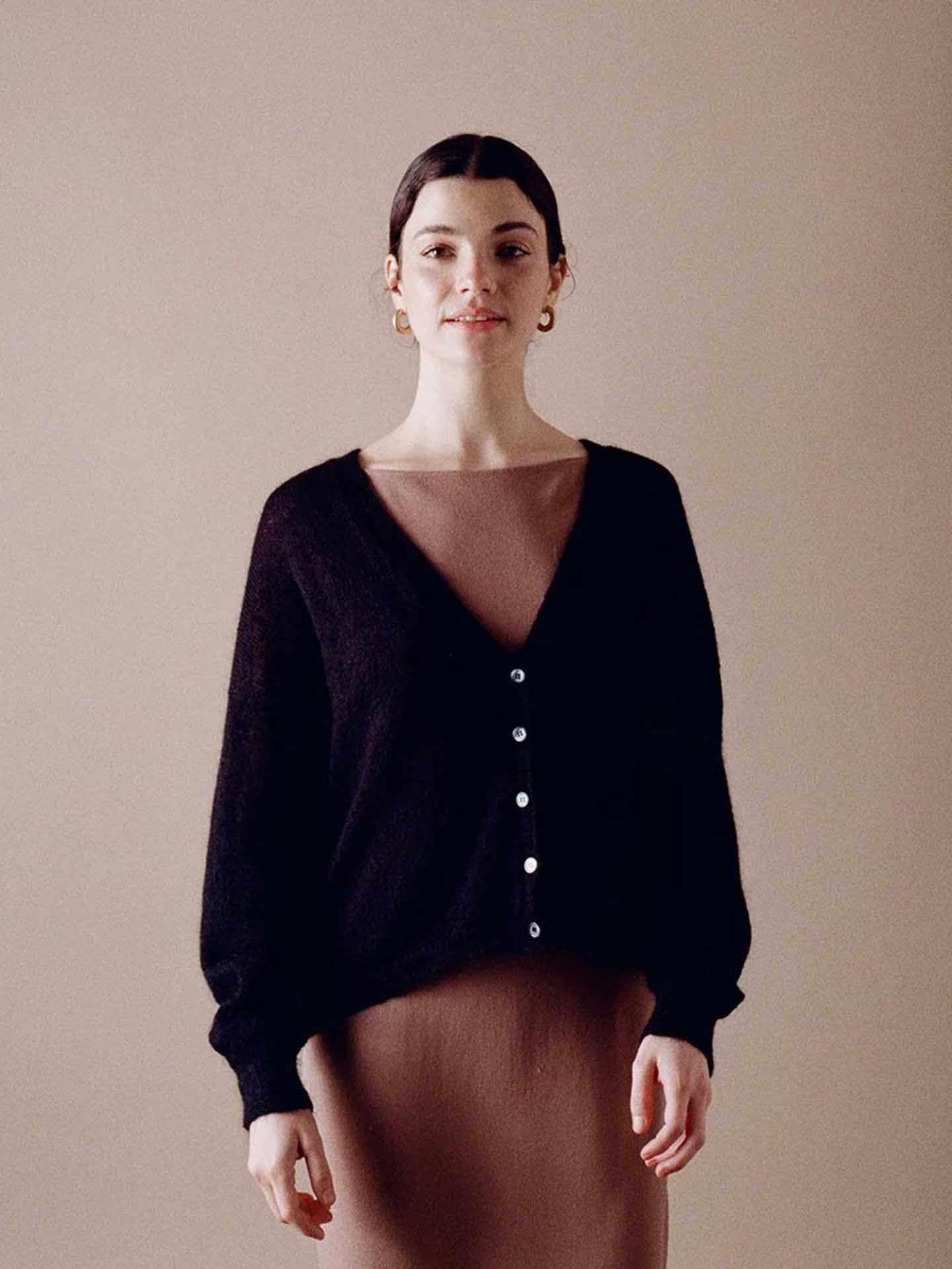 A petite person stands against a beige background, wearing the Francie Mist Cardigan in black over a brown dress.