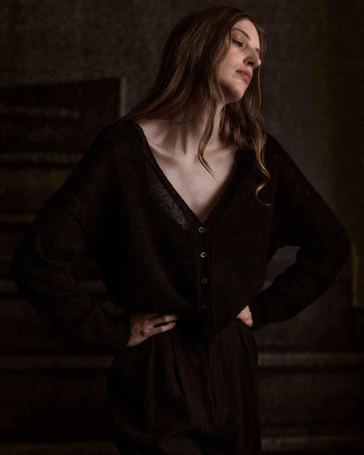 A petite woman with long hair stands confidently, her hands on her hips in a relaxed pose. She wears the Francie Mist Cardigan in black against the dimly lit background.