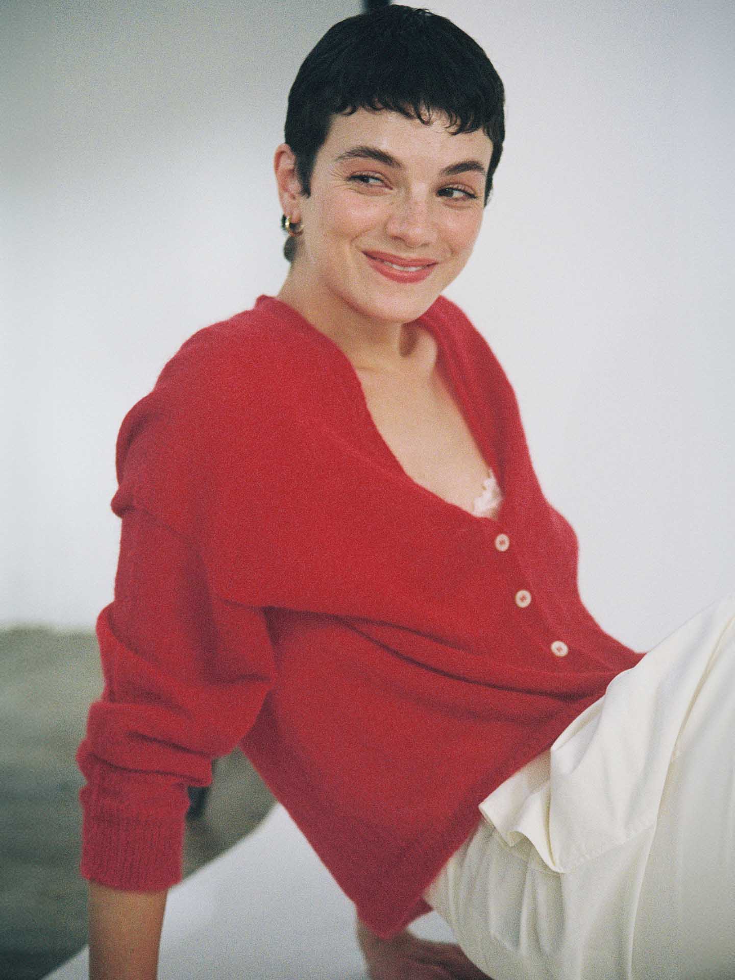 A petite person wearing the Francie Mist Cardigan in Cherry, a relaxed, cropped style, paired with white pants, sits and smiles slightly against a neutral background.