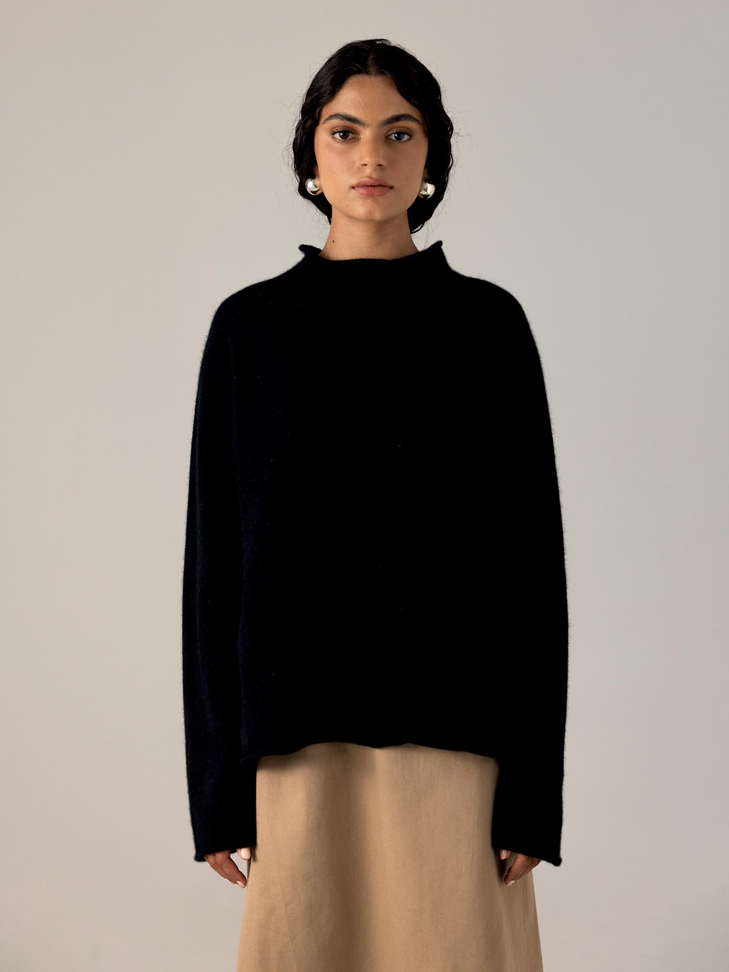 A petite woman with dark hair in a Francie Eclipse Knit – Black sweater and beige skirt stands against a neutral background, looking directly at the camera.