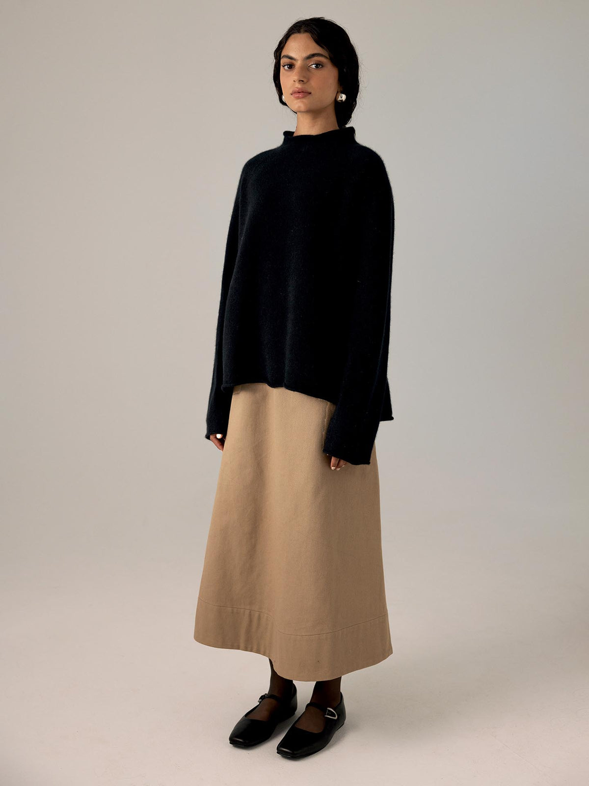 A petite woman in a Francie black Eclipse Knit sweater and beige skirt stands on a light gray background. She is wearing black shoes and looking at the camera.