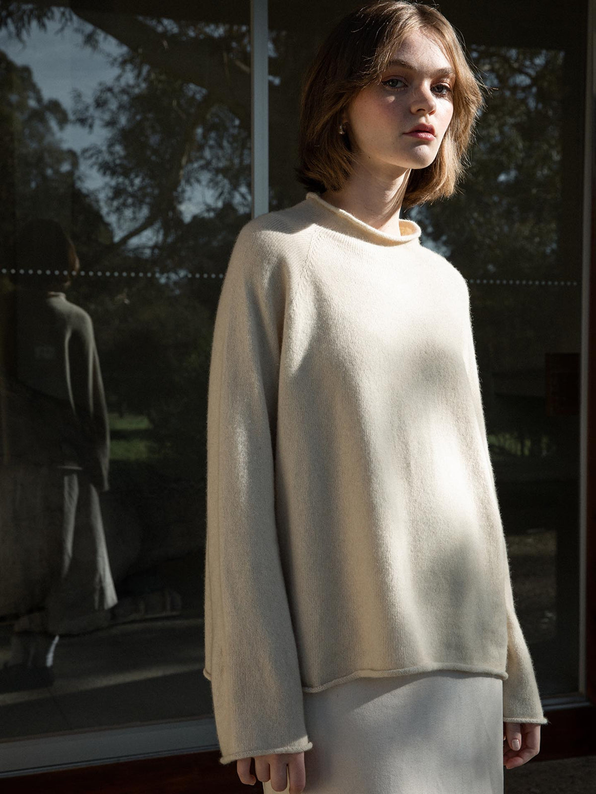 Petite young woman in an oversized Francie white sweater standing in sunlight by a window, with her reflection visible on the glass.