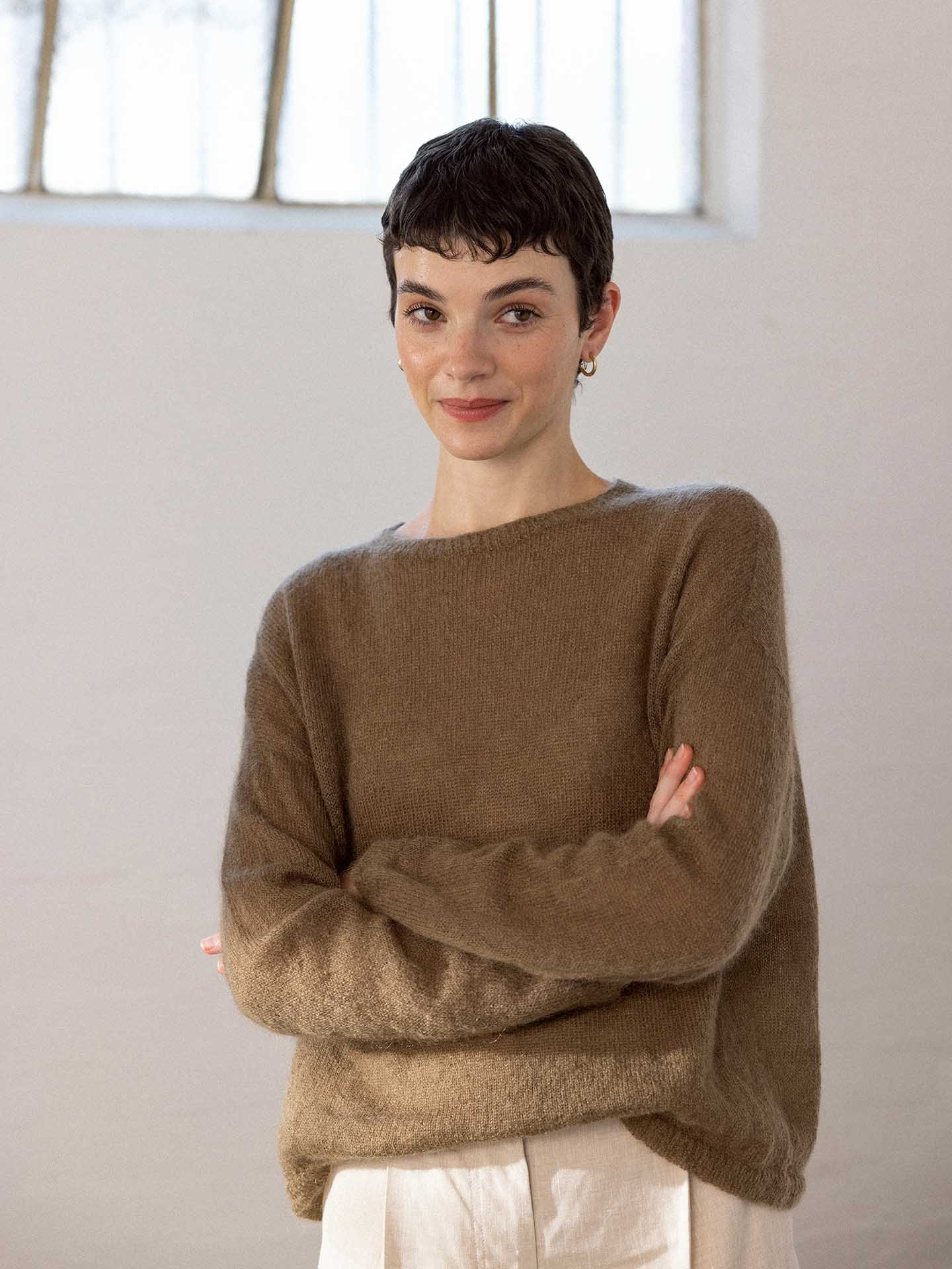 A petite person with short dark hair wears the oversized "Feather Knit – Hazel" sweater by Francie and white pants, standing with arms crossed in a softly lit, minimalistic room.