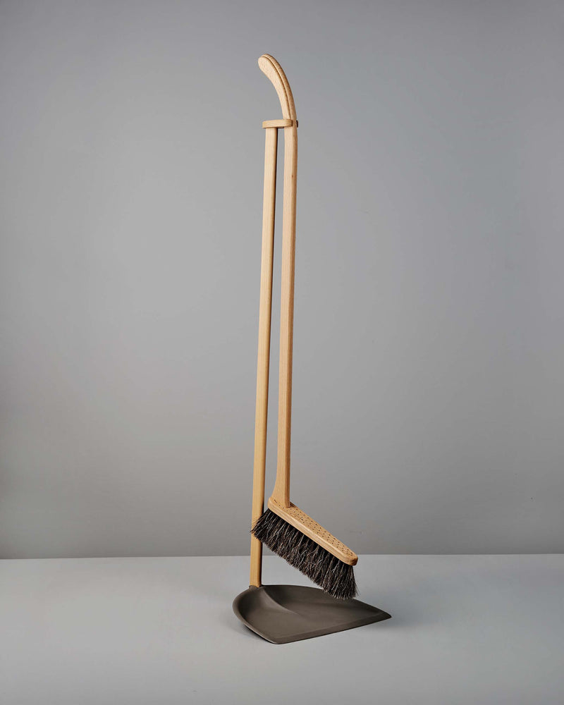 A wooden-handled broom with dark bristles is resting upright on a metal dustpan against a plain gray background.