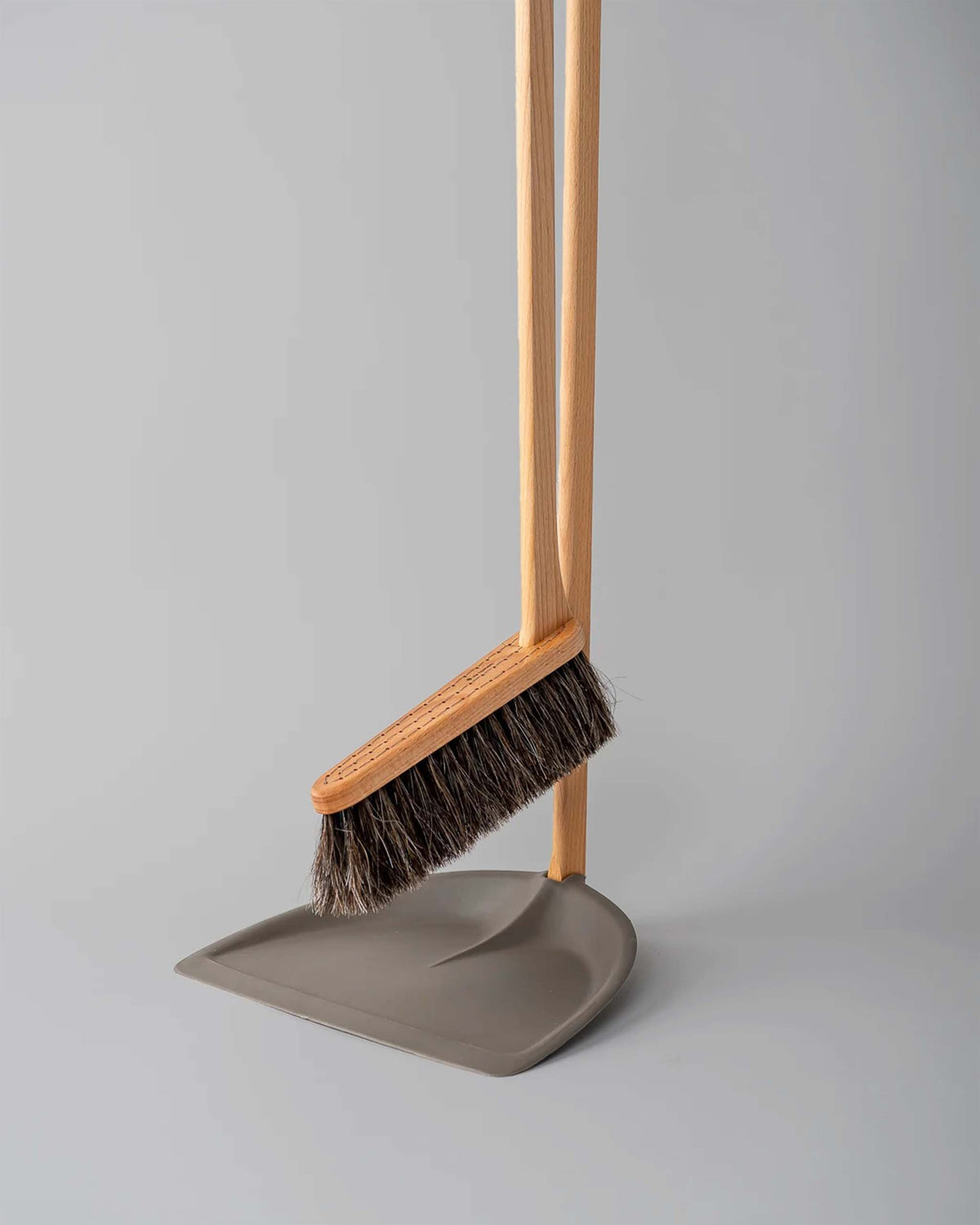 A wooden broom with dark bristles rests on a grey dustpan, both standing upright against a plain grey background.
