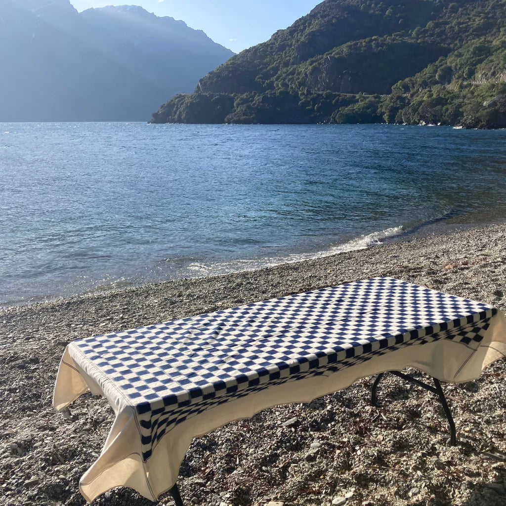 A table adorned with the Stitchwallah Ikat Weave Tablecloth – Indigo Check is set up on a pebbled shore near a body of water, with mountains in the background.