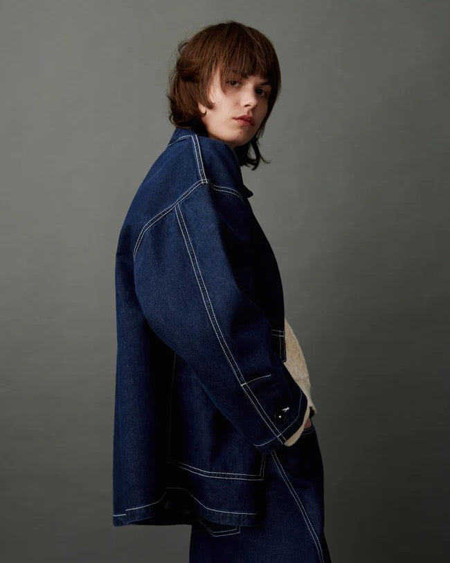 A person with medium-length brown hair wears the Portrait Jacket – Indigo with matching dark denim pants, standing against a plain gray background.