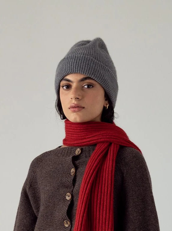 A person wearing the Hinoki Beanie – Granite, a brown buttoned sweater, and a red scarf stands against a plain light background.