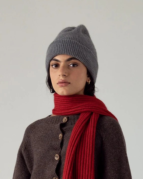 A person wearing the Hinoki Beanie – Granite, a brown buttoned sweater, and a red scarf stands against a plain light background.