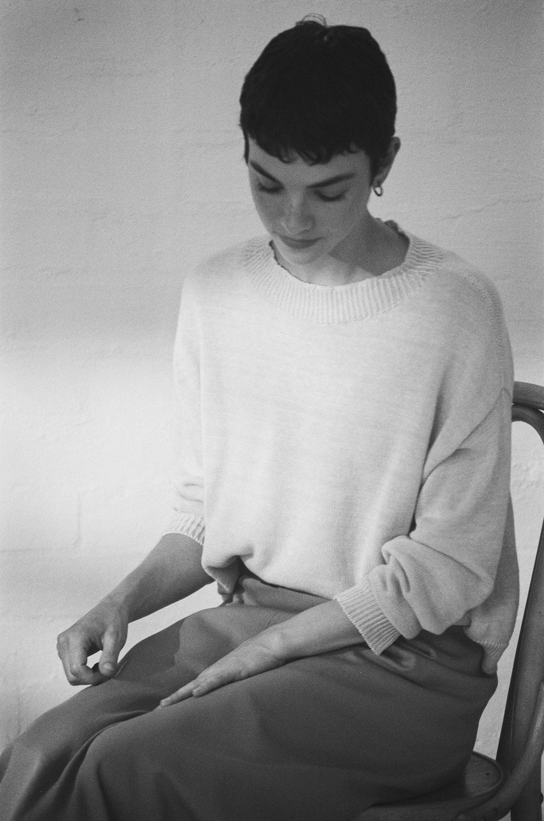 Seated on a chair in a softly lit room, a person with short hair and hands on their lap wears the Pebble Knit – Creme sweater in a relaxed fit, paired with a long skirt.