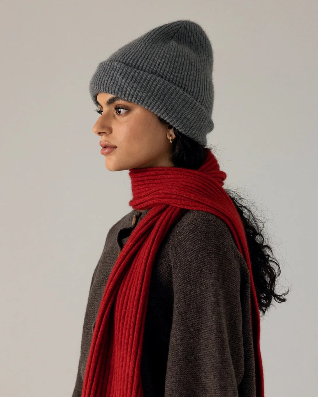Person in profile view wearing the Hinoki Beanie – Granite, a brown sweater, and a long red scarf, standing against a plain light background.