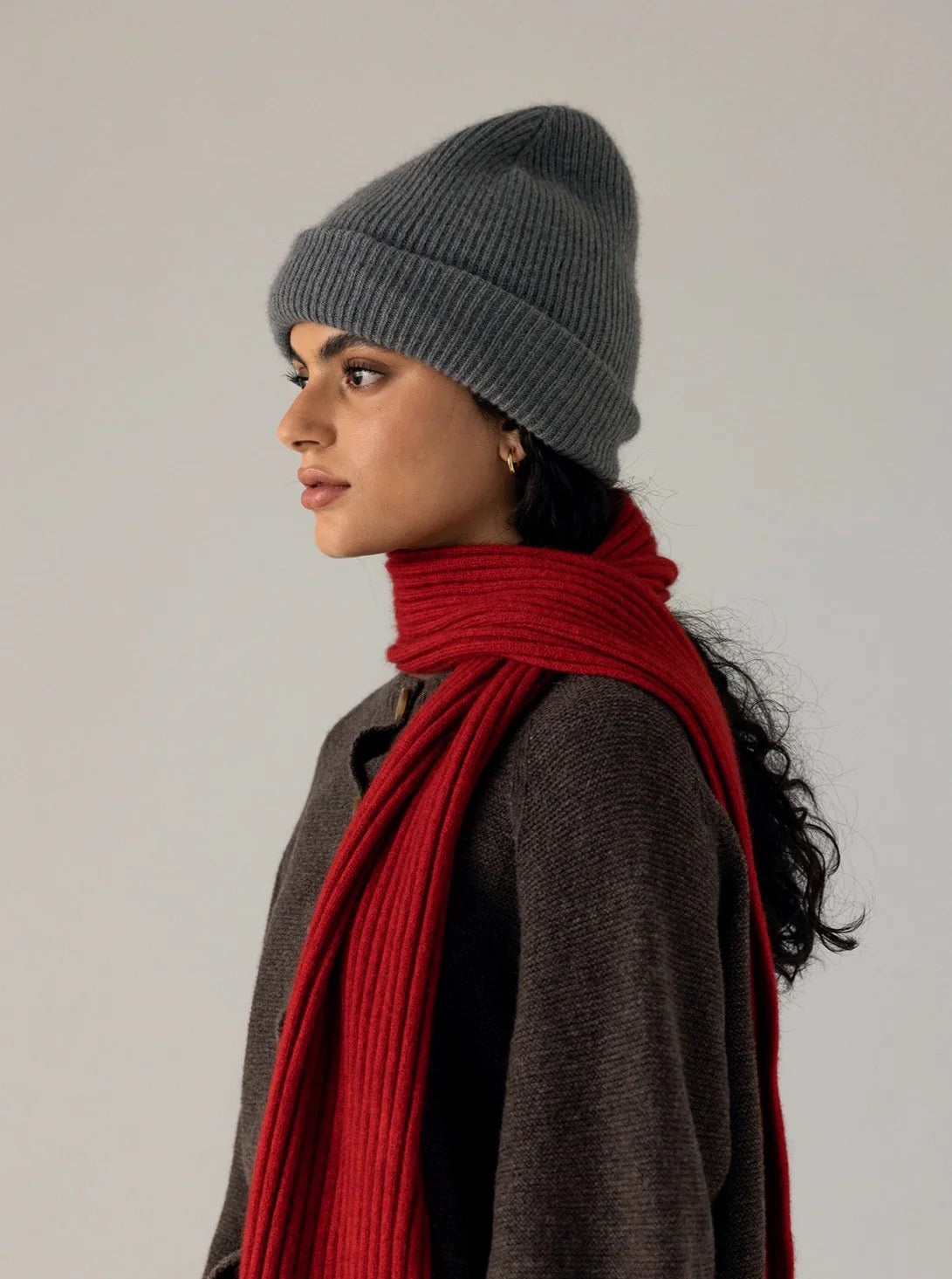 Person in profile view wearing the Hinoki Beanie – Granite, a brown sweater, and a long red scarf, standing against a plain light background.