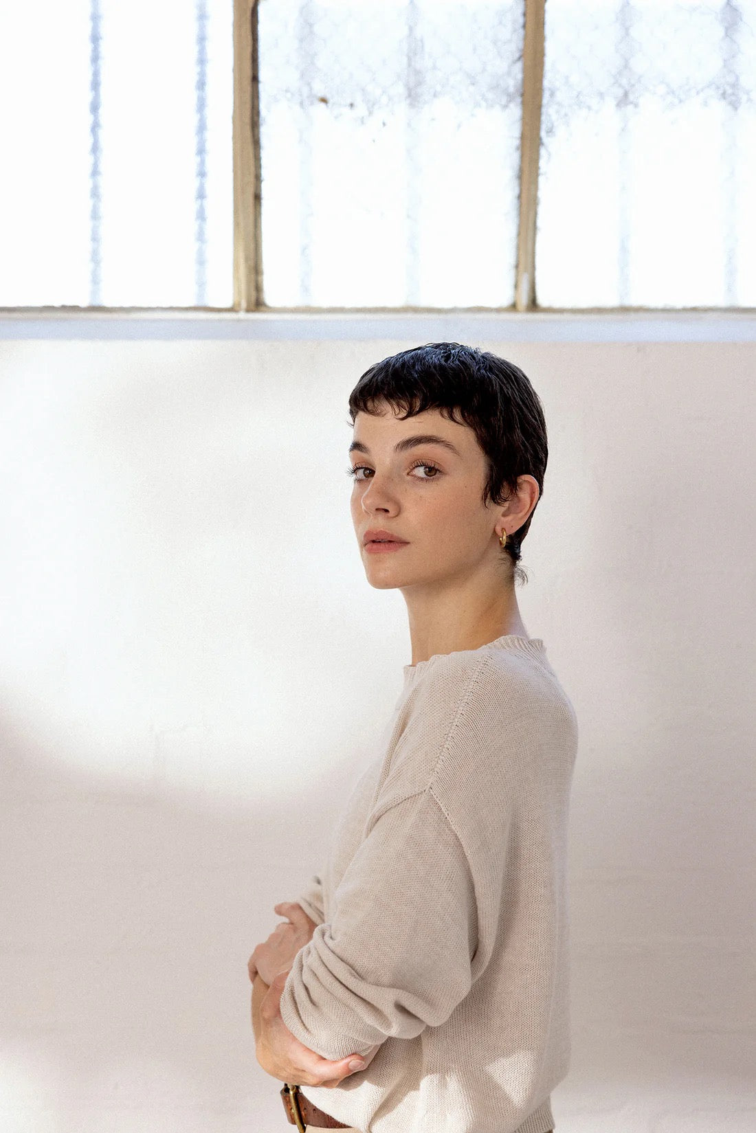 A person with short dark hair, wearing Pebble Knit – Creme in a relaxed fit, stands indoors near a bright window, facing slightly sideways with arms crossed.