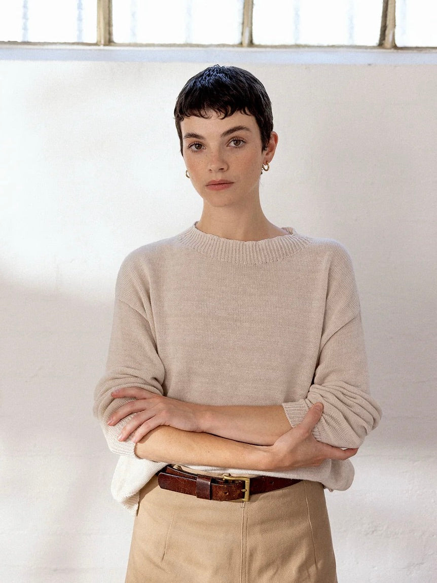A petite person with short dark hair poses indoors in front of a white wall, wearing the Pebble Knit – Creme sweater styled cropped with beige pants, a brown belt, and hoop earrings, arms crossed in a relaxed manner.