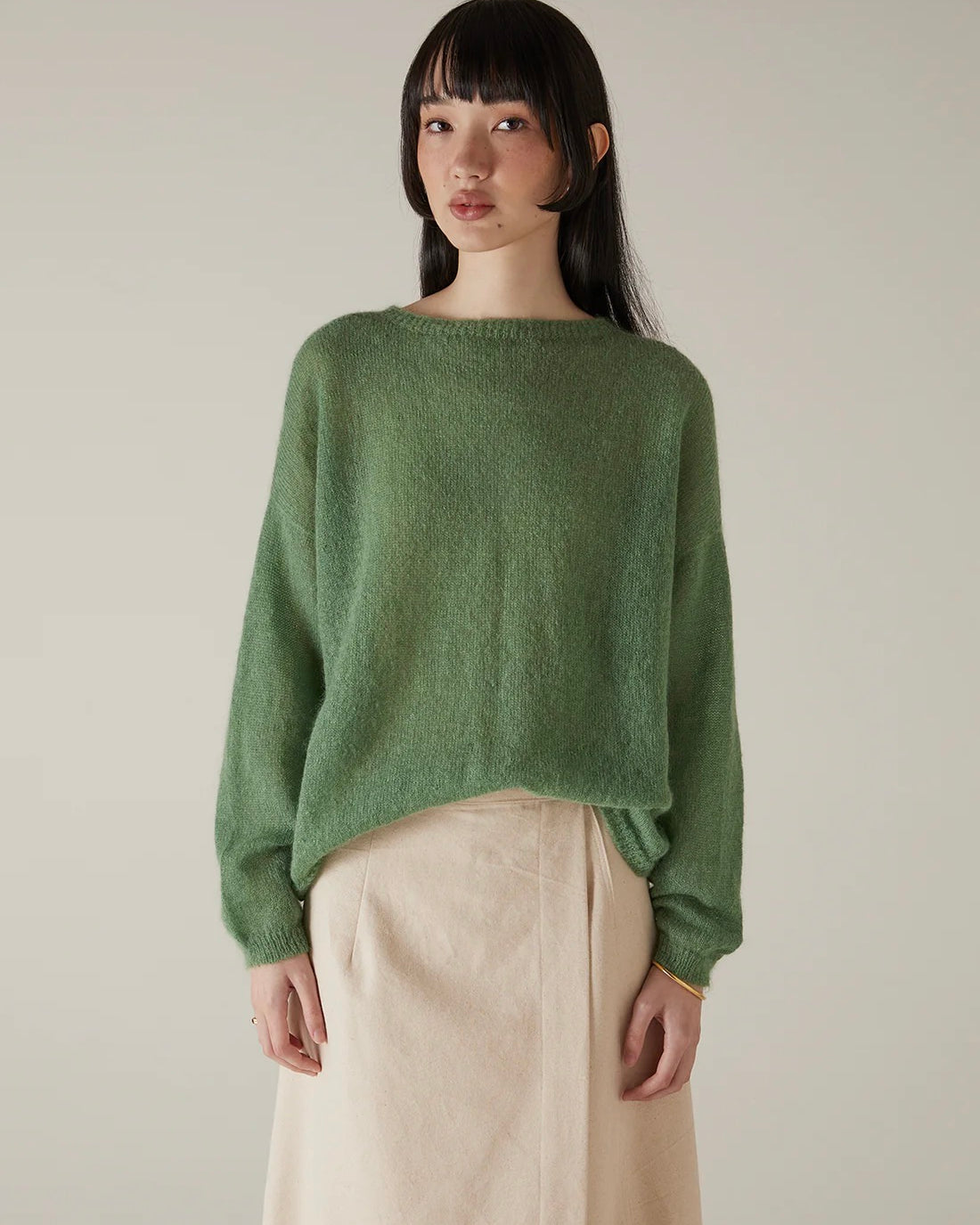 A woman stands against a plain background, wearing the Feather Knit – Clover in a relaxed fit with a light beige skirt, looking directly at the camera with a neutral expression.