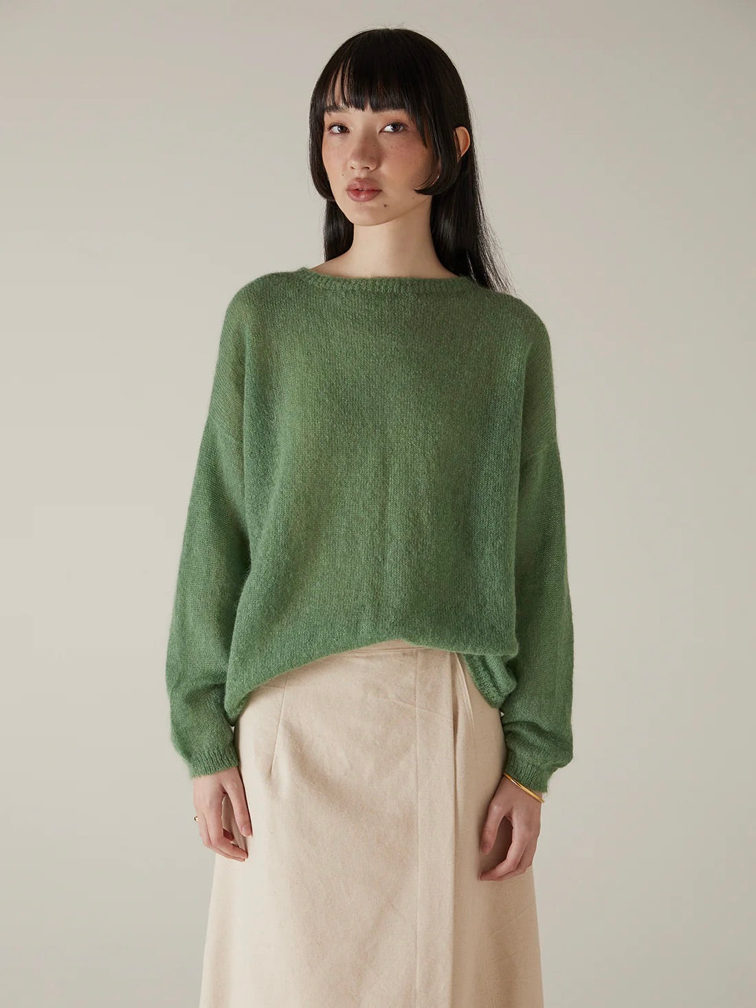 A woman stands against a plain background, wearing the Feather Knit – Clover in a relaxed fit with a light beige skirt, looking directly at the camera with a neutral expression.