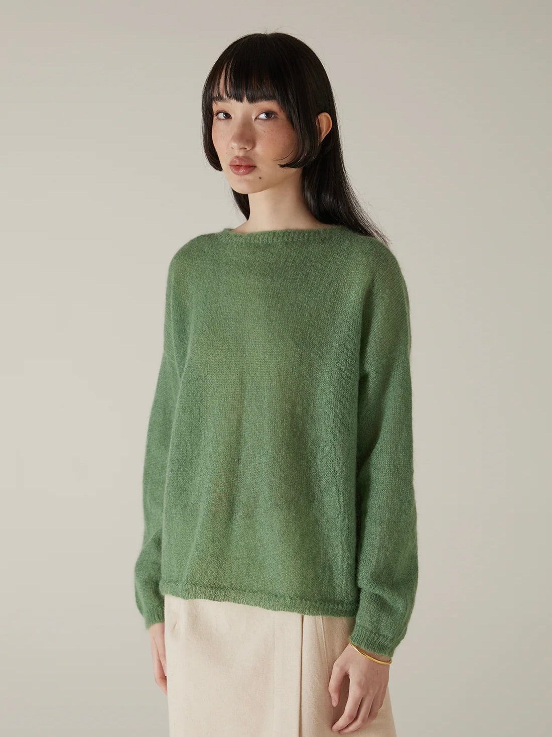 A person with straight dark hair wears the Feather Knit – Clover, a relaxed fit green sweater, paired with beige pants, standing against a plain light background.